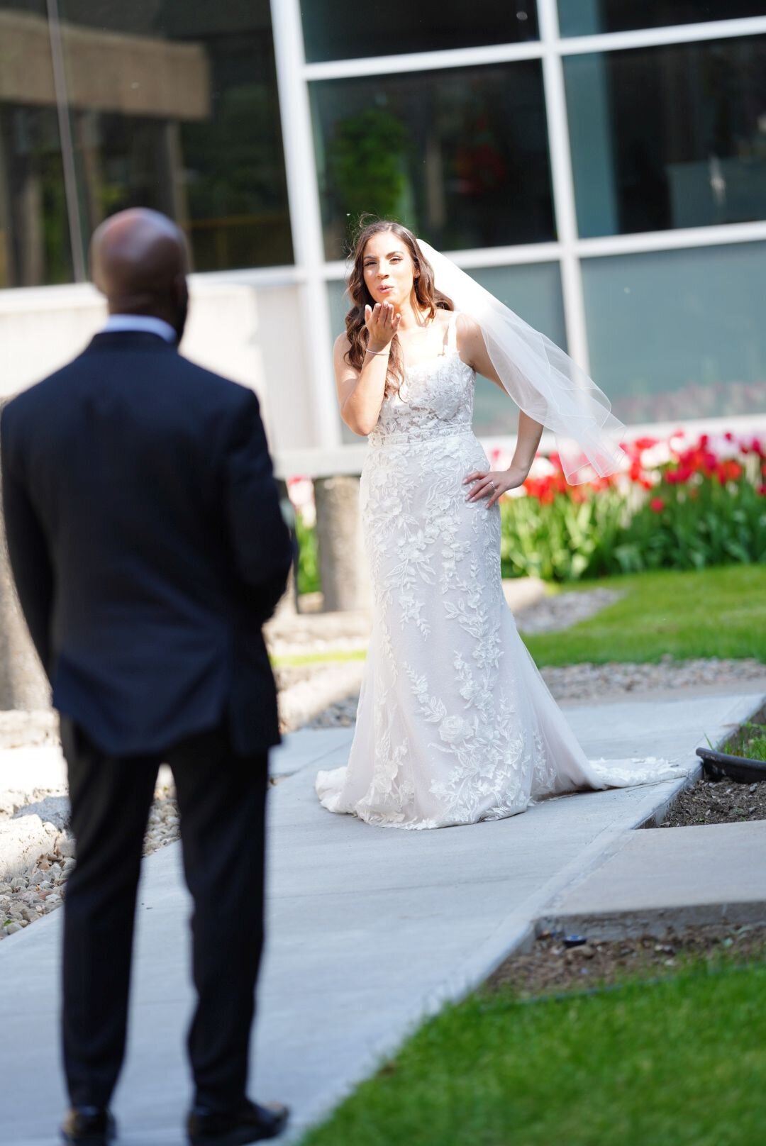 A playful moment captured from behind the groom of the bride as she blows a kiss to the groom, who stands a short distance away. The image highlights their fun-loving relationship and adds a touch of whimsy to the wedding day memories.