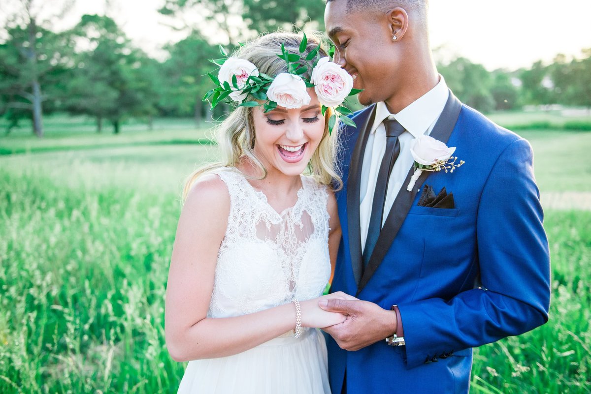 Couple holding hands outside of a country club with field behind them, she's wearing a flower crown and laughing and he has a blue tux on