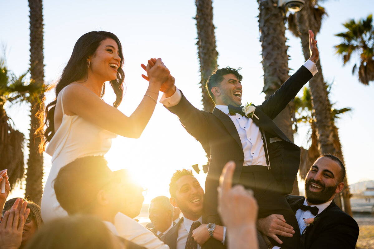 Professional San Francisco Bay Area wedding photography capturing authentic moments as bride in white dress and groom in black tuxedo celebrate with raised hands in Santa Monica with tropical palm trees and warm golden sunset creating joyful atmosphere