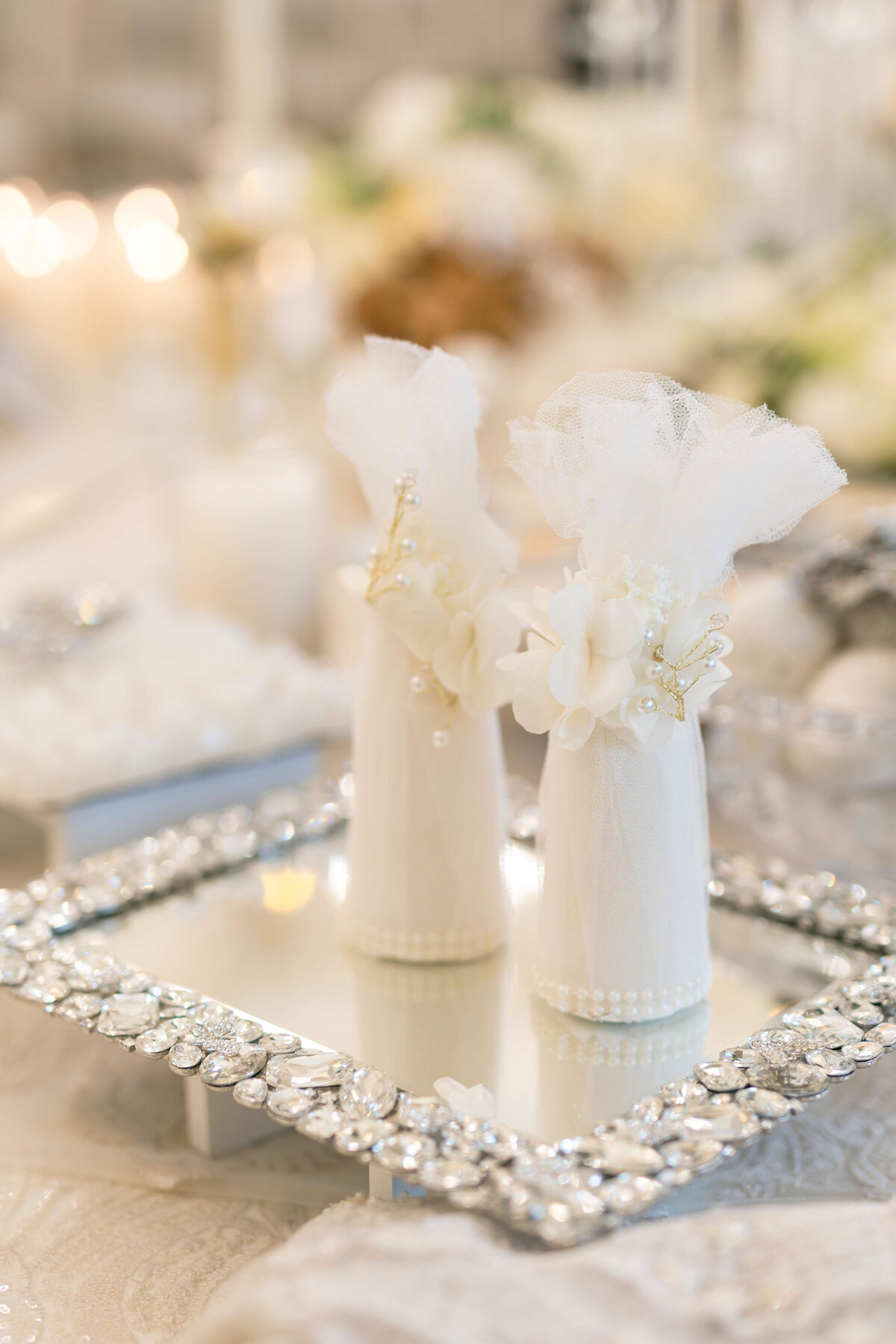 Elegant wedding table setting with two small white tulle-wrapped gifts or favors on a mirrored tray, decorated with pearls and flowers. The background is softly blurred, highlighting the sparkling and delicate details of the arrangement.