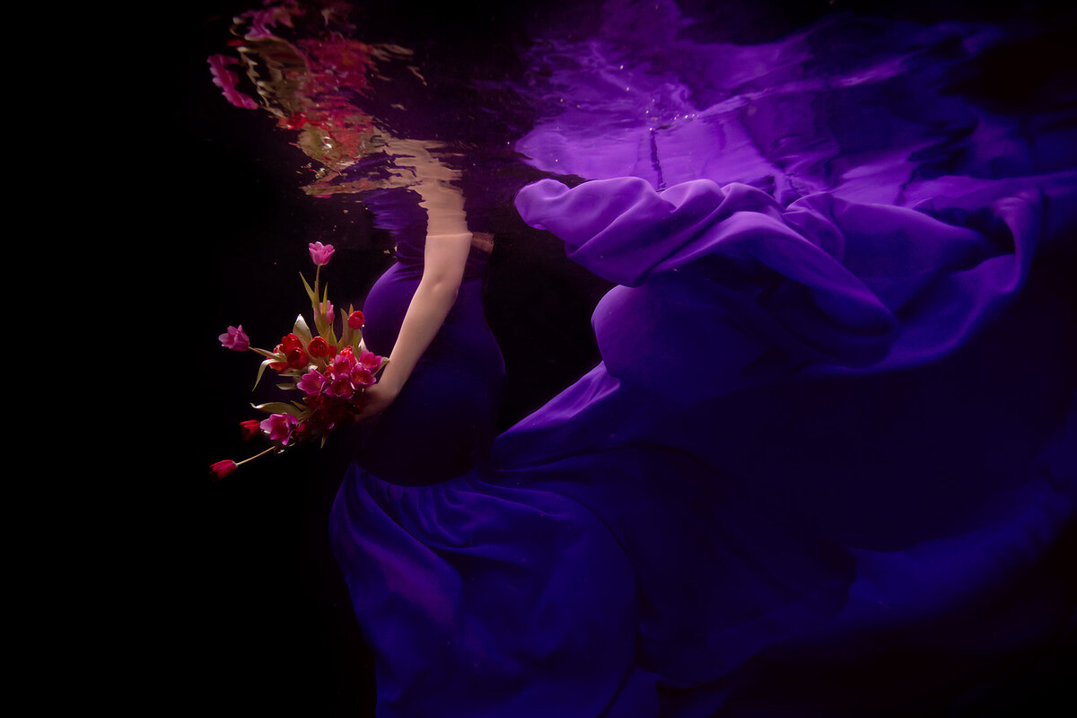 An ethereal underwater maternity portrait featuring a woman gracefully holding a bouquet of vivid pink tulips. The flowing purple gown surrounds her in a dreamy display of elegance and serenity.