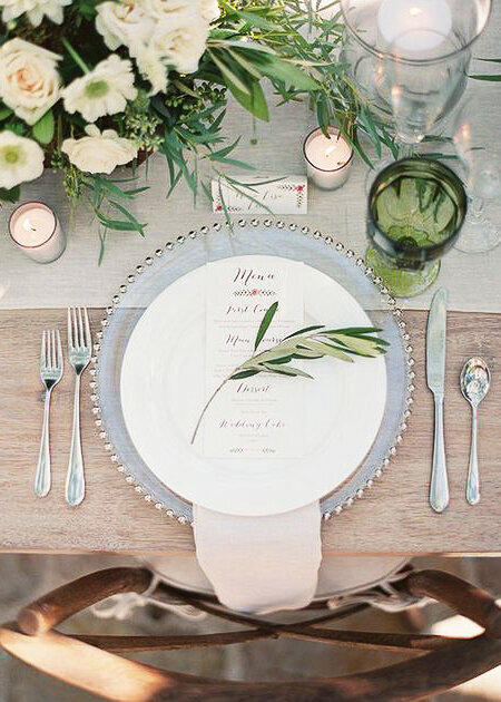 A wedding place setting with an olive twig over a white menu with green glassware and flowers.