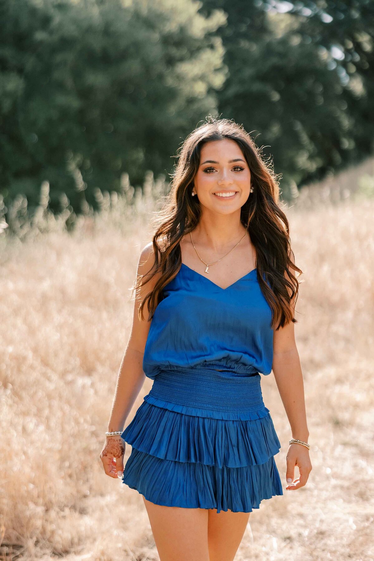 Brunette girl wearing a royal blue dress and looking at the camera while walking through tall, dry grass