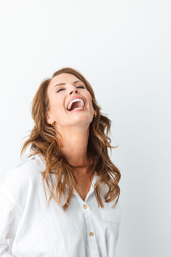 Girl in white on a white backdrop throws her head back and laughs.