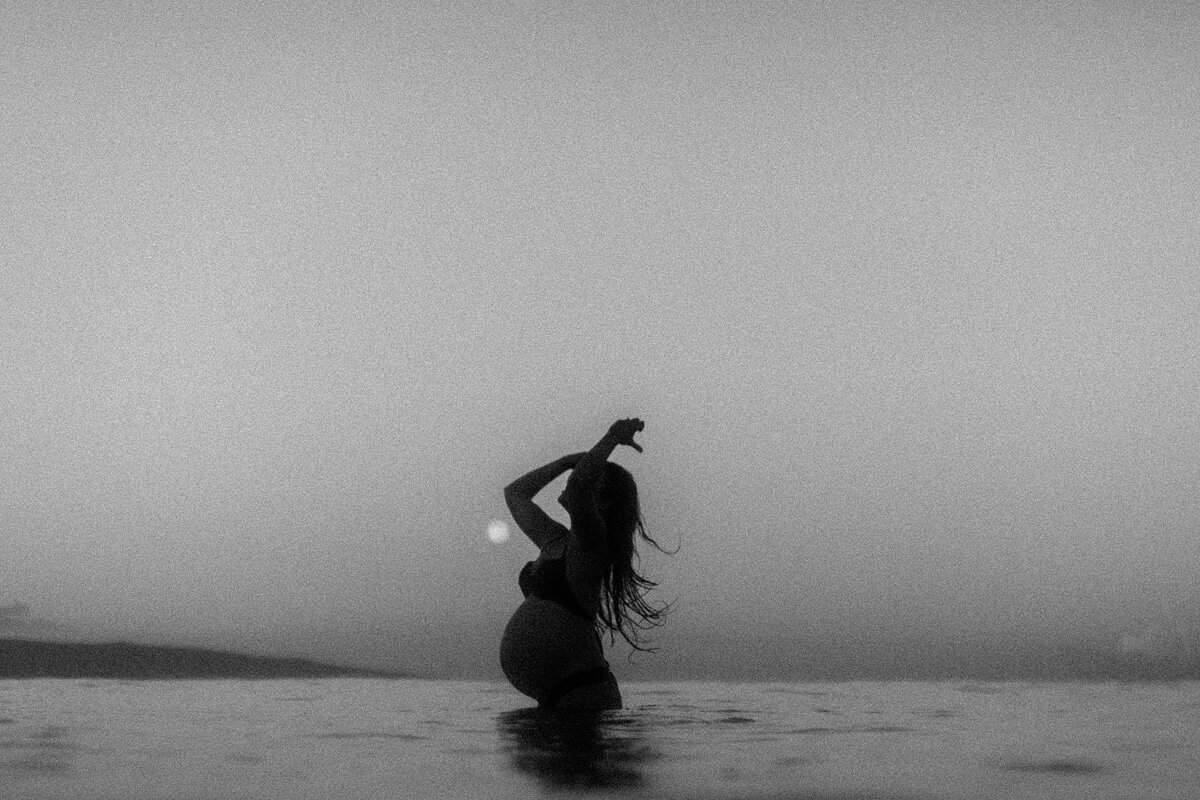 Powerful black and white Maui babymoon portrait feautring woman tossing hair back int the water at sunrise.