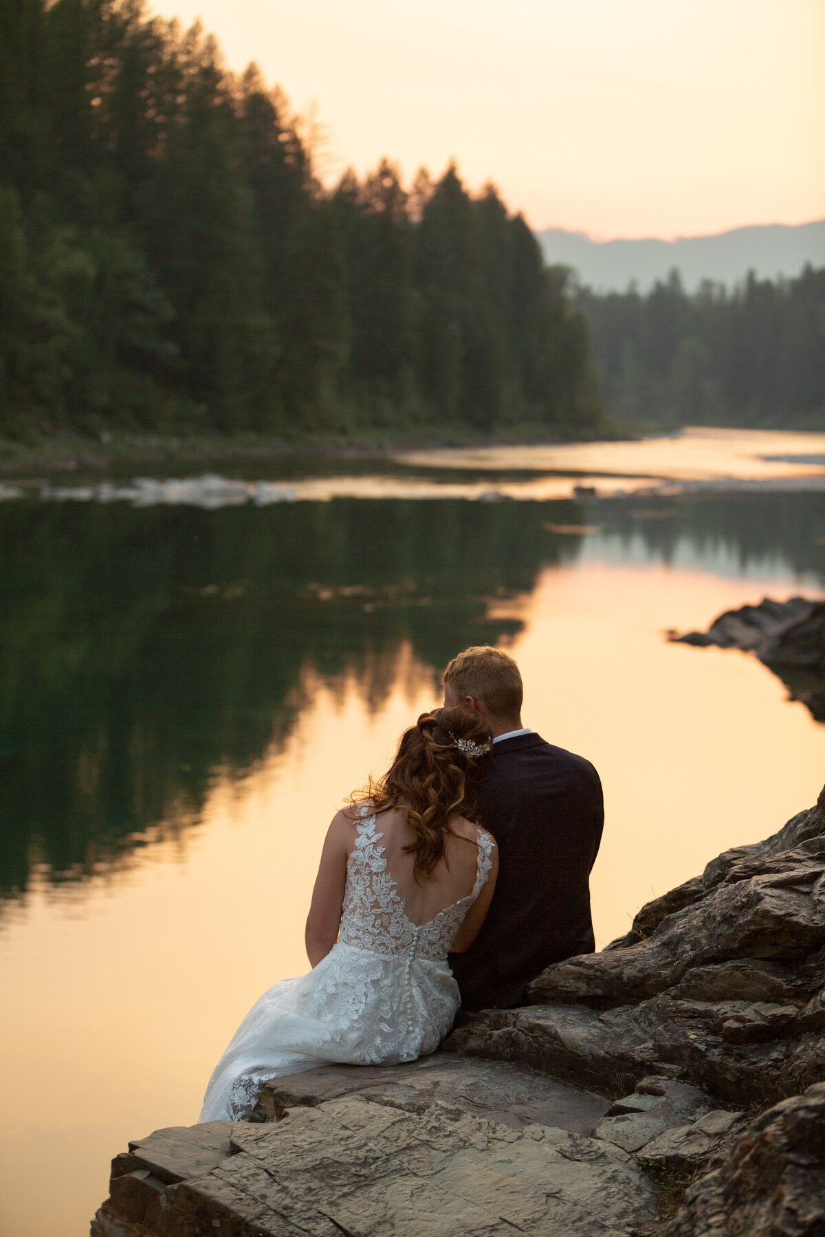A couple sit on a rock overlooking a river as the sunsets and casts an orange glow around them in Glacier National Park.