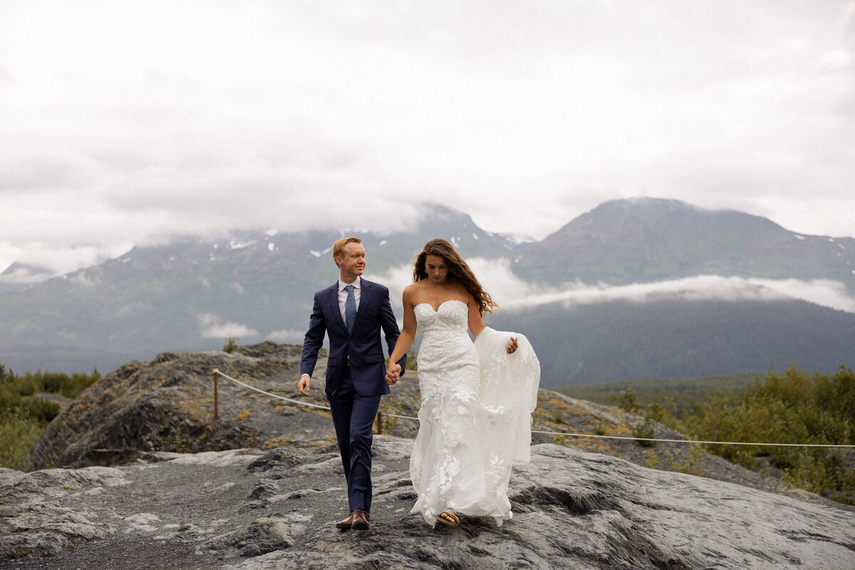 A wedding couple holding hands and walking along a rocky mountainside 