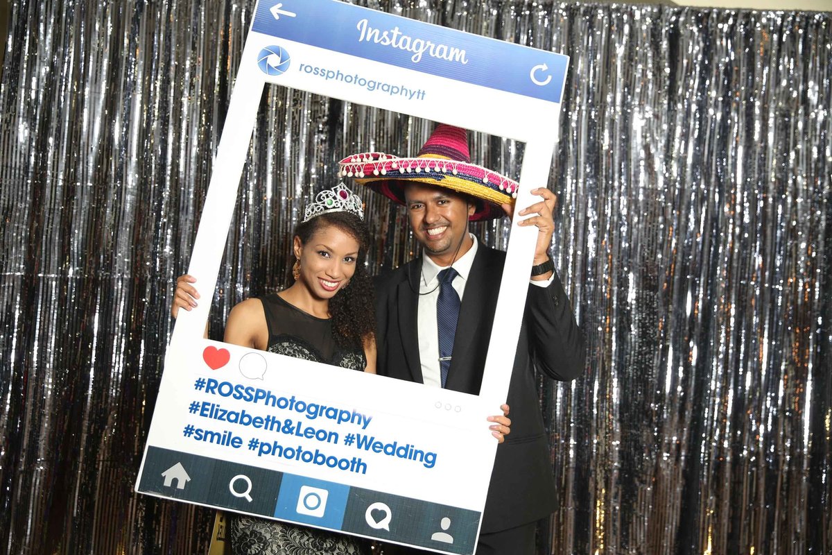 Couple poses in front of silver backdrop with customized, social media cut-out frame. Photobooth by Ross Photography, Trinidad, W.I..