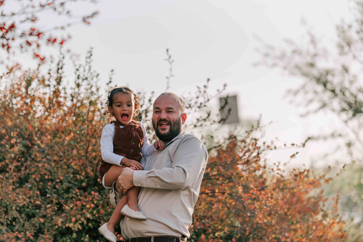 Dad hold littke girl in front a fall bush. 