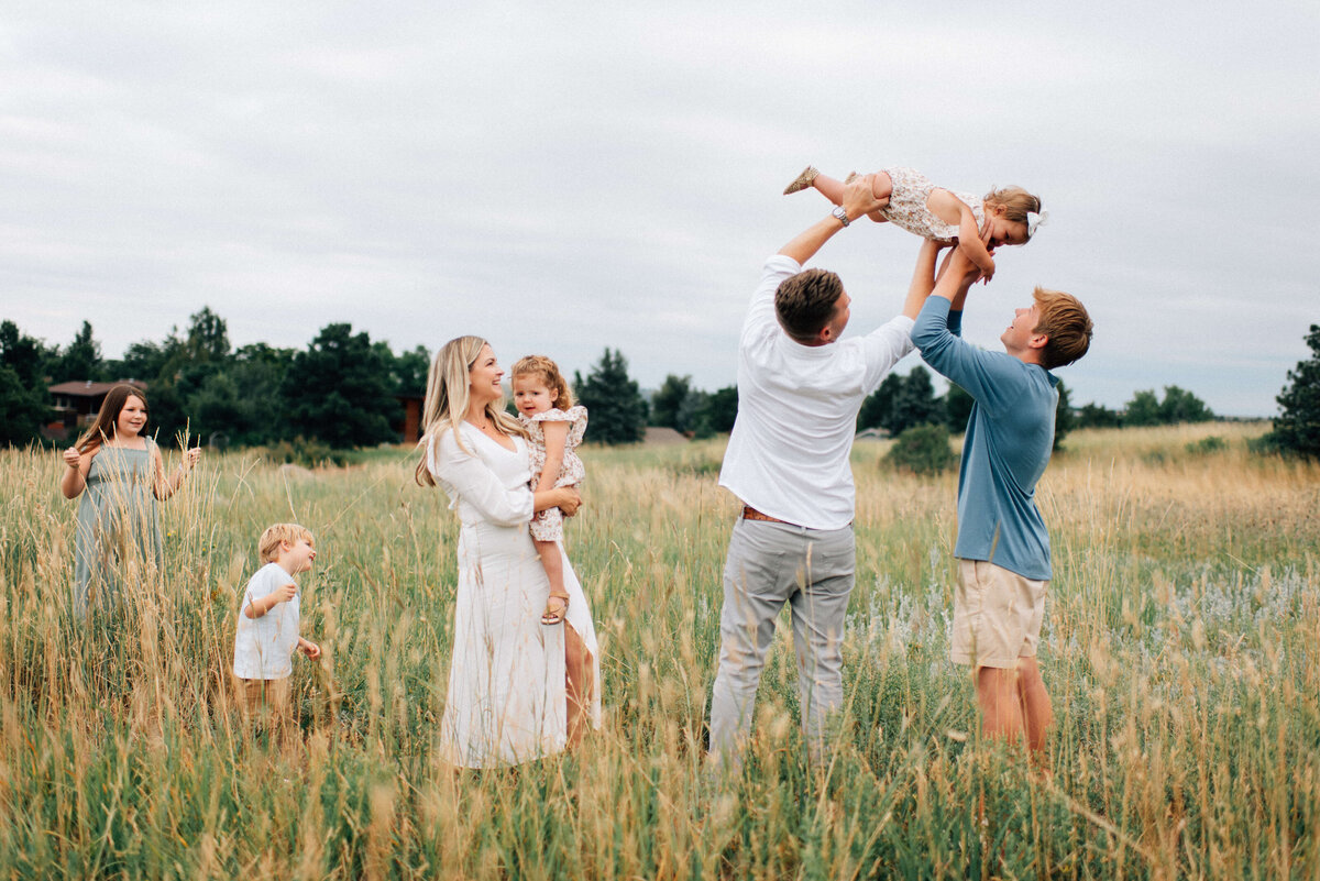 large family of seven playing in the field with the youngest baby girl getting lifted to the sky by her dad and big brother