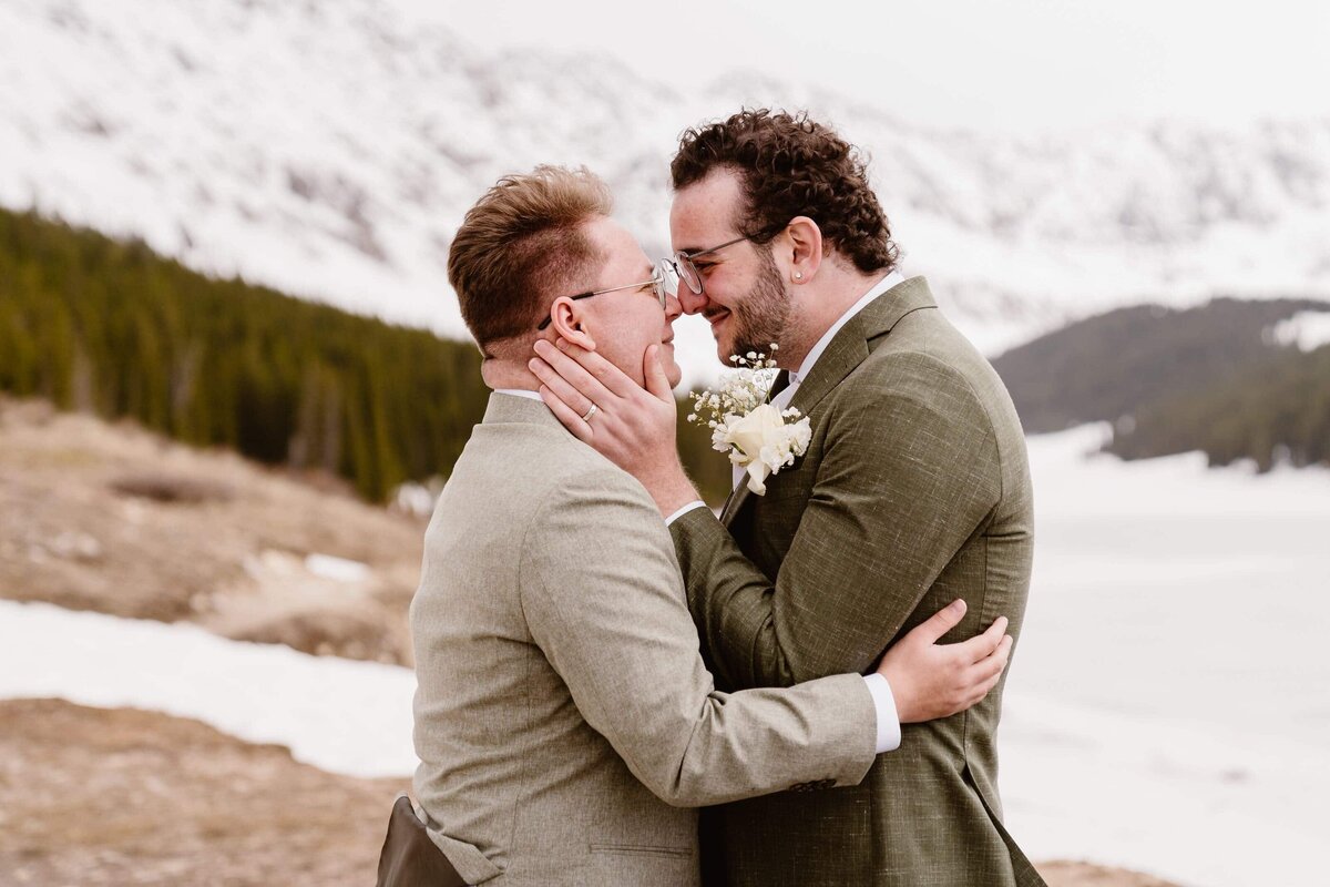 Two grooms embrace and smile with snowy mountain peaks behind them during their Breckenridge, Colorado elopement.