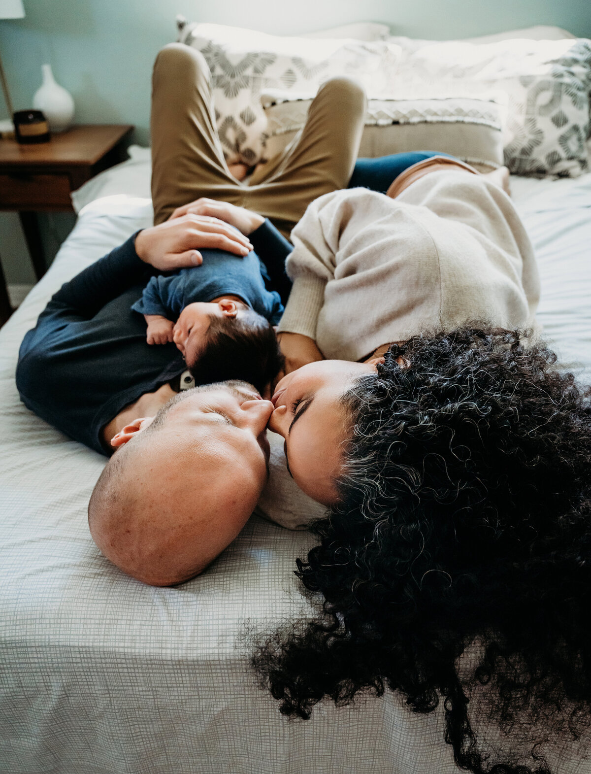 Newborn Photography, dad lays on the bed with baby, mom gives him a kiss beside him