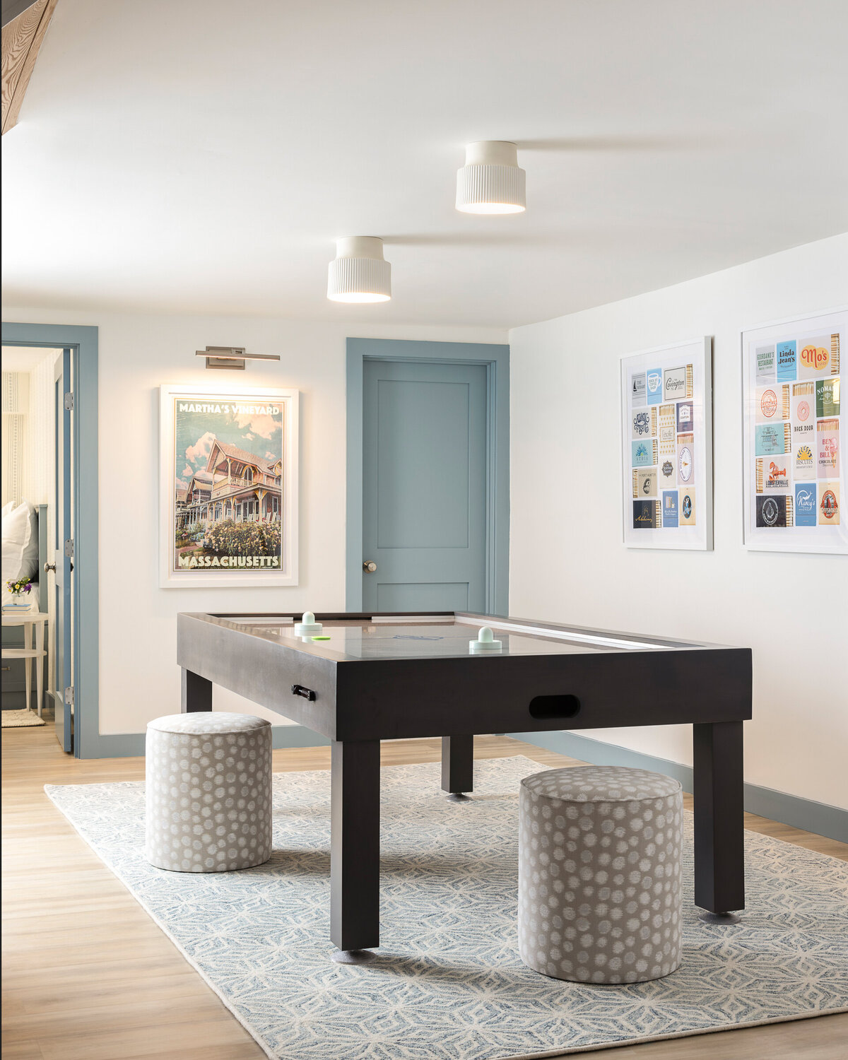 Bright game room featuring a black air hockey table, two patterned round stools, light blue doors, framed art on the walls, and ceiling-mounted lights.