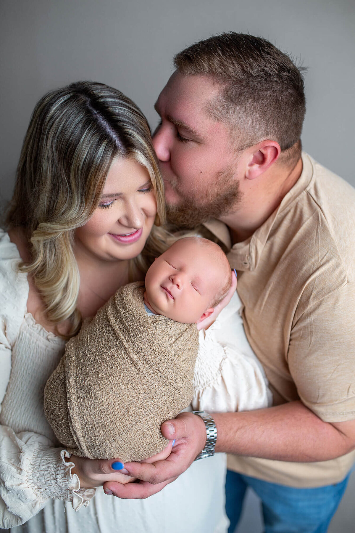 Man kissing woman while she looks lovingly at newborn baby boy wrapped in neutral tones 