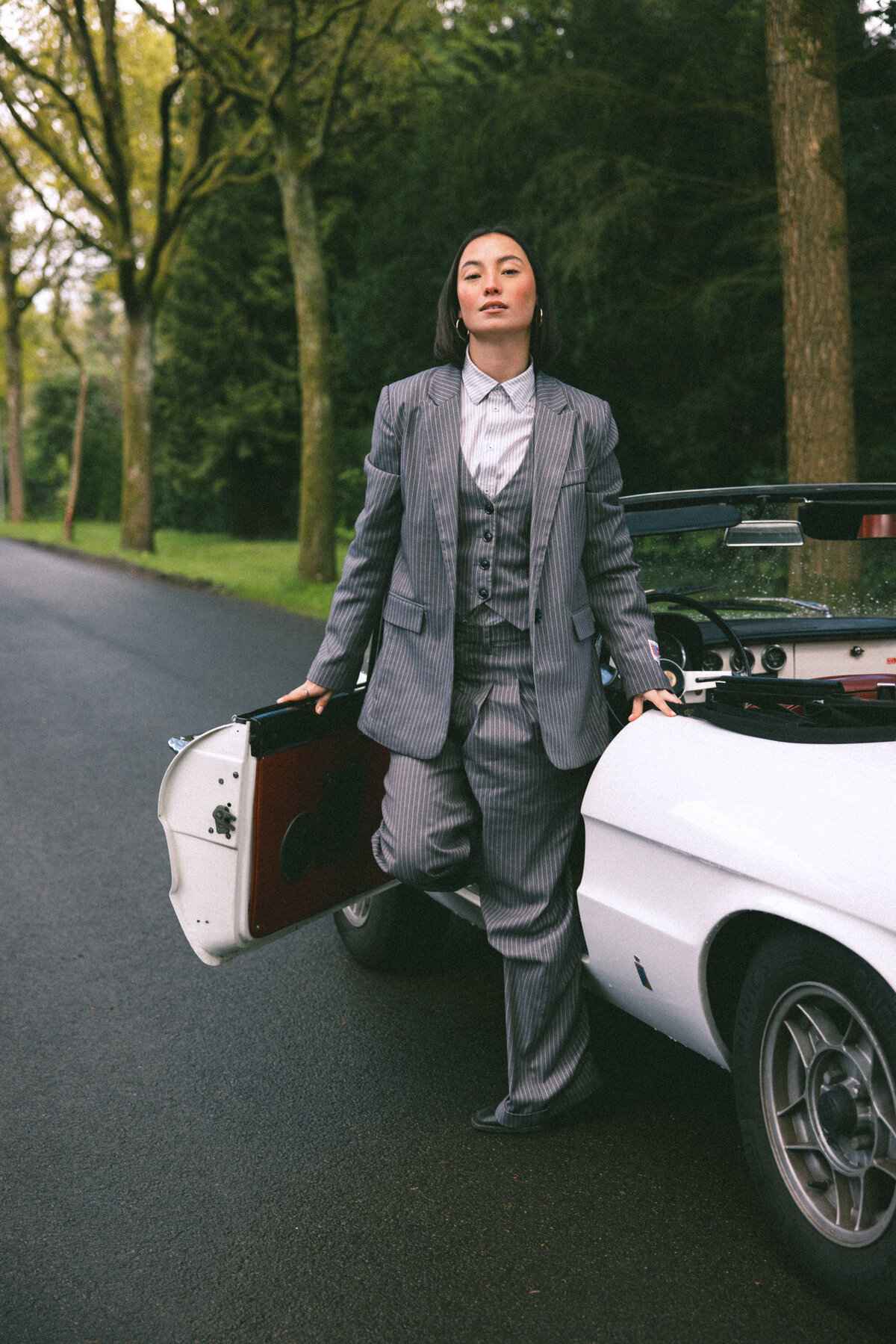 A captivating rear view of the subject walking away from the camera, towards the open road. Her tailored suit and the classic car parked beside her create a narrative of adventure and independence, enhanced by the natural surroundings, photographed by Bo Eppink.