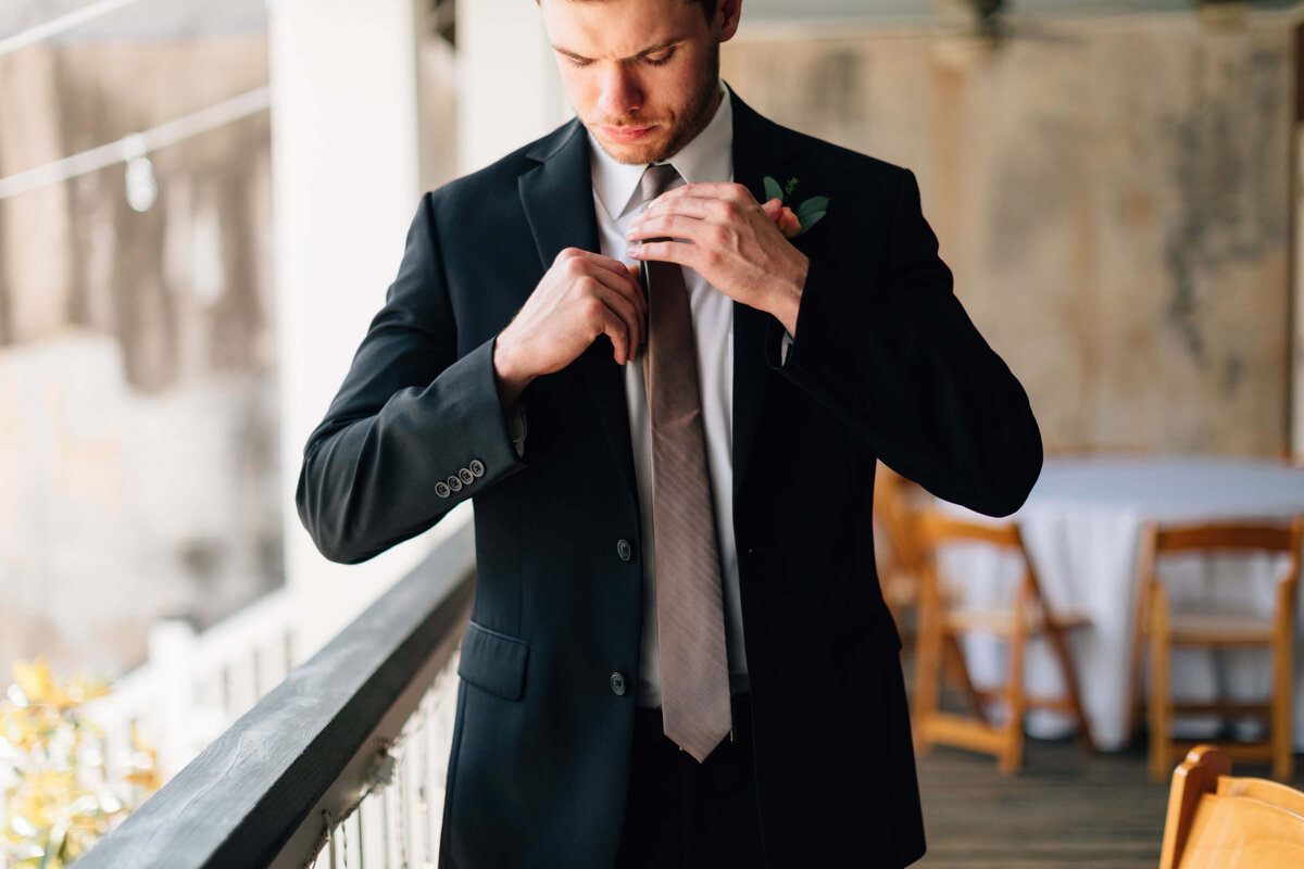 A man in a black suit adjusts his light-colored tie while standing on a balcony. In the background, there are wooden chairs and a round table with a white tablecloth.