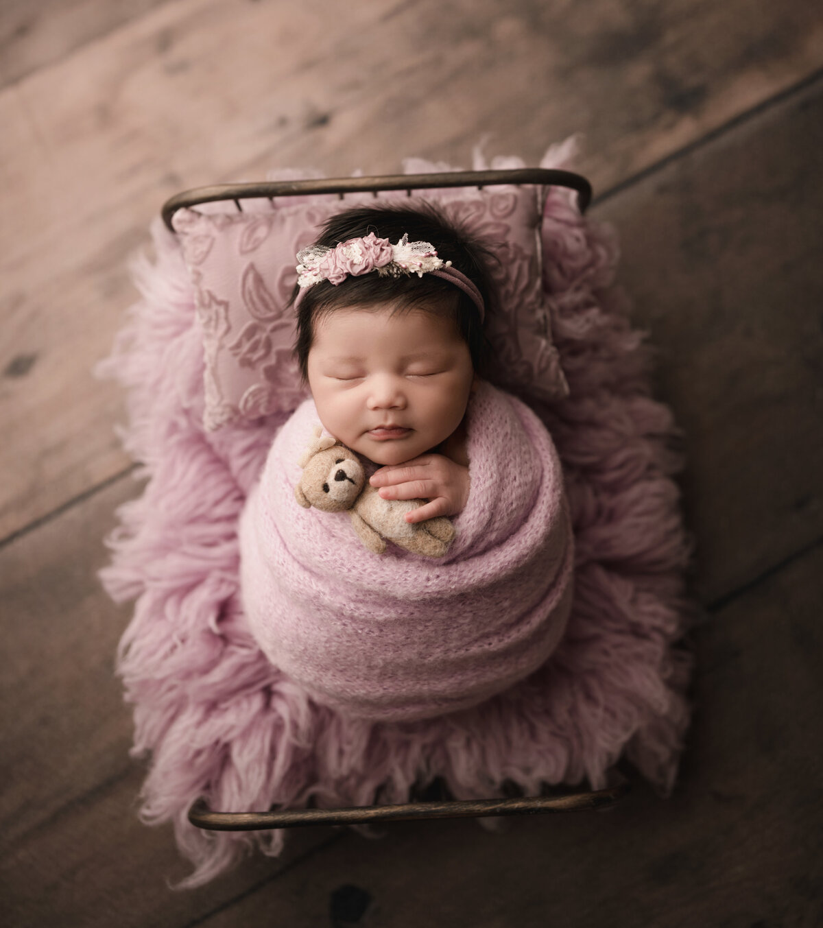 Baby girl is wrapped in a pink knit swaddle and placed atop of a miniature newborn prop bed for her Riverside, CA newborn photoshoot. Baby is sleeping an her hand is peeking out of the swaddle and holding a miniature felt teddy bear. Captured by best Riverside newborn photographer Bonny Lynn Photography.
