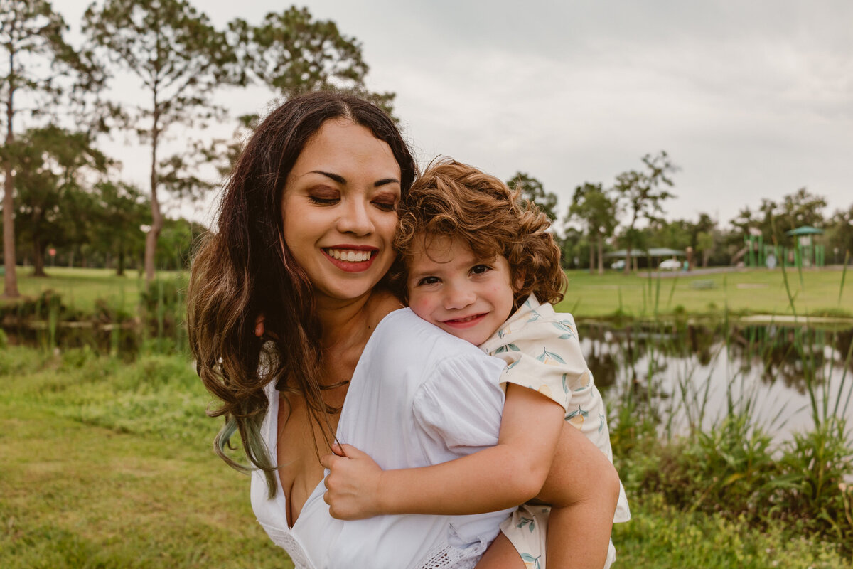 Happy mother carrying her young son on her back at Walter Hall Park in League City, Texas. Both are smiling with greenery and a serene park backdrop. Captured by ShutterUp Photography.