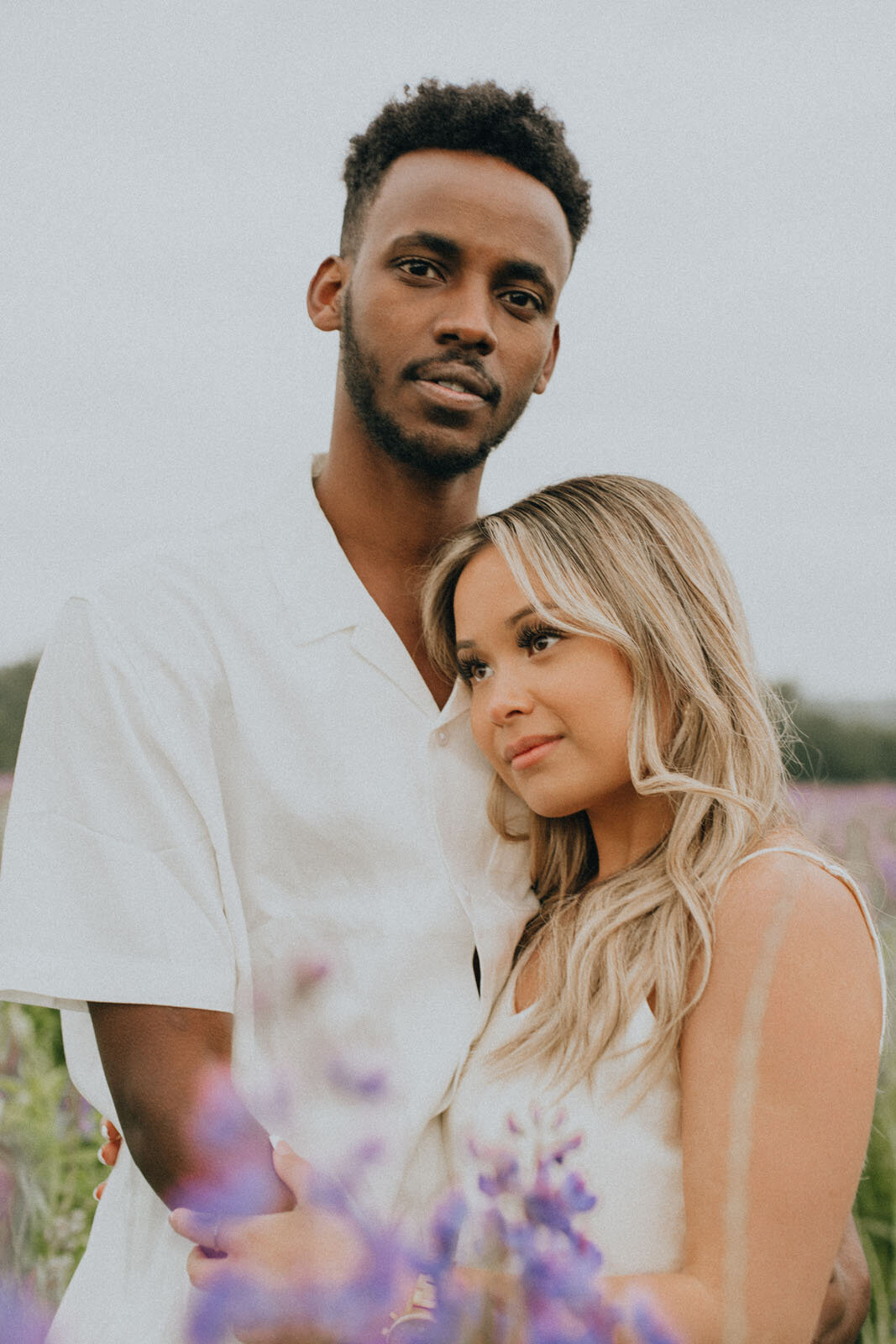 Girl leaning on guy's chest while guy looks into the camera in a lupine field