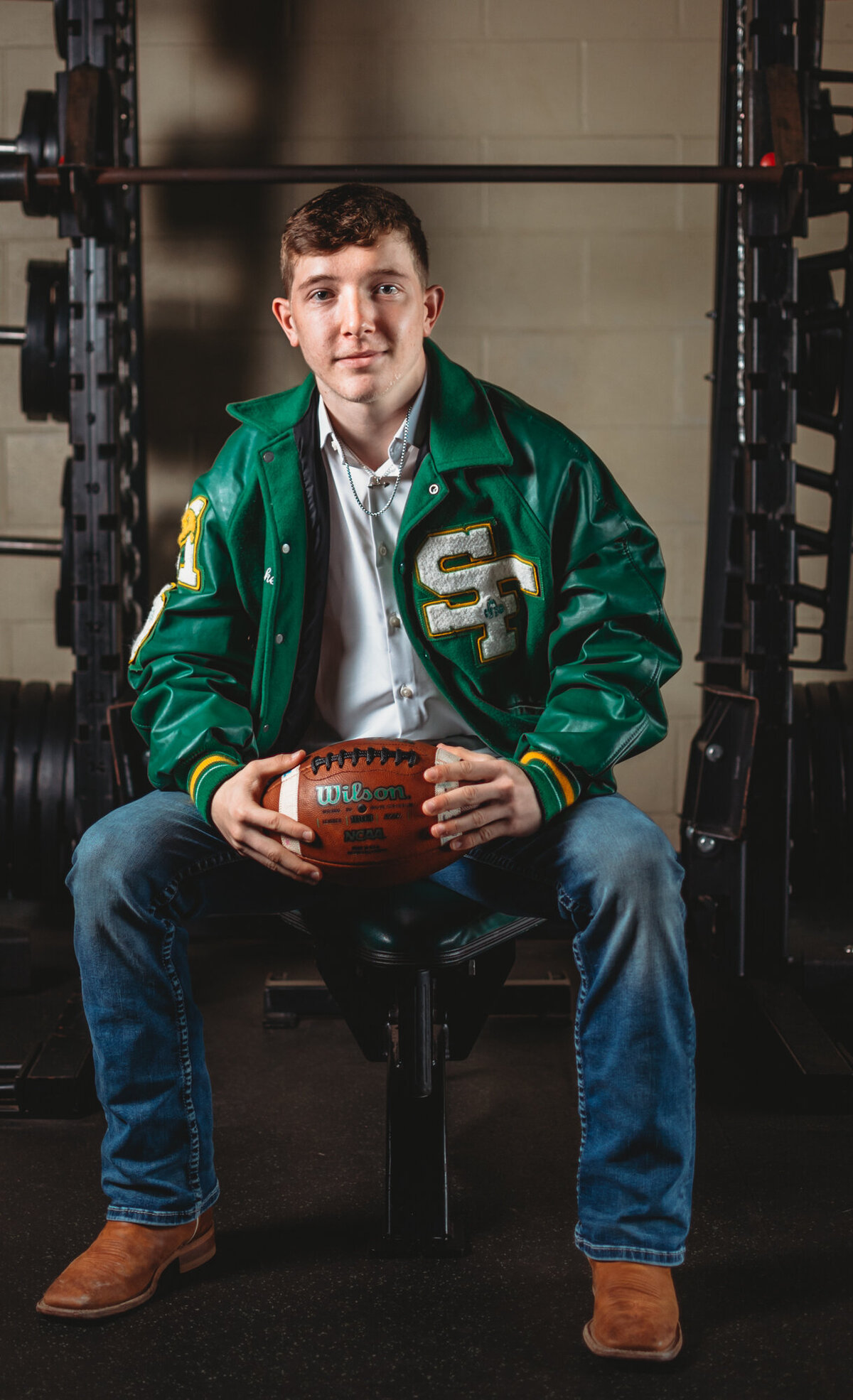 A recent graduate sits on a weightlifting bench and holds a football .