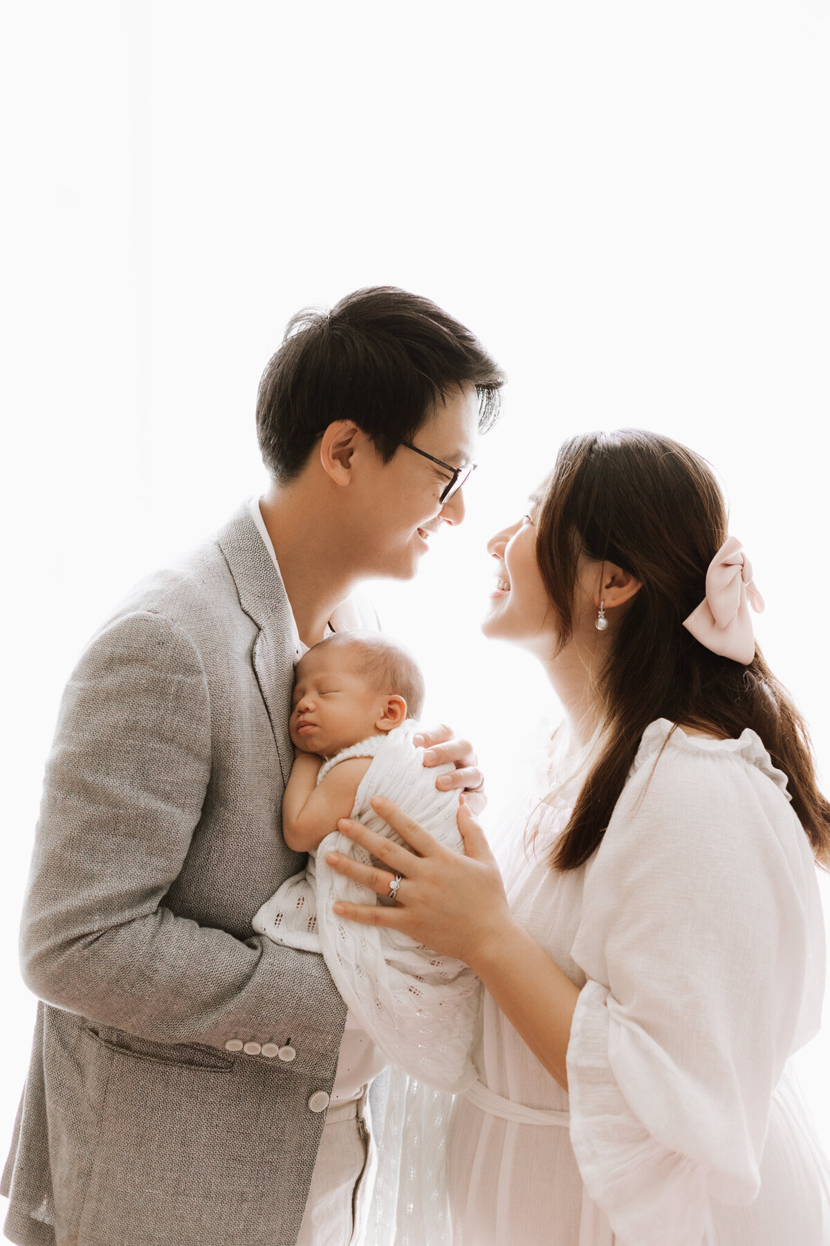 A joyful young couple holds their sleeping newborn baby during a studio session. The father, dressed in a grey suit, cradles the baby close to his chest, while the mother, wearing a flowing white dress and pink bow in her hair, smiles up at him. Captured in a softly lit Sydney newborn photography studio, this image radiates warmth, connection, and the tender joy of early parenthood.