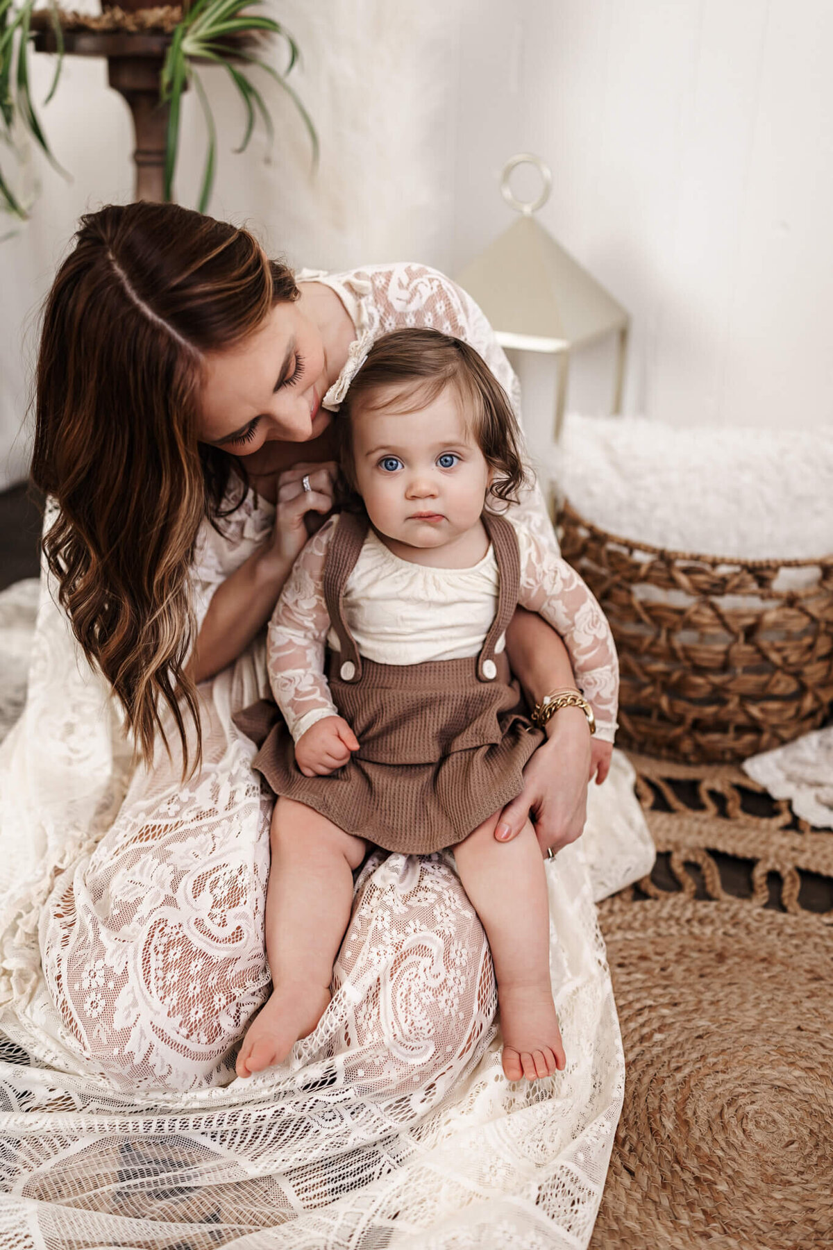 A woman with long brown hair, wearing a lace dress, lovingly cradles a baby on her lap. The baby, dressed in a brown outfit and boasting blue eyes, gazes around as they sit on a woven rug. Captured by a Bloomington family photographer, this moment radiates warmth in the cozy ambiance.