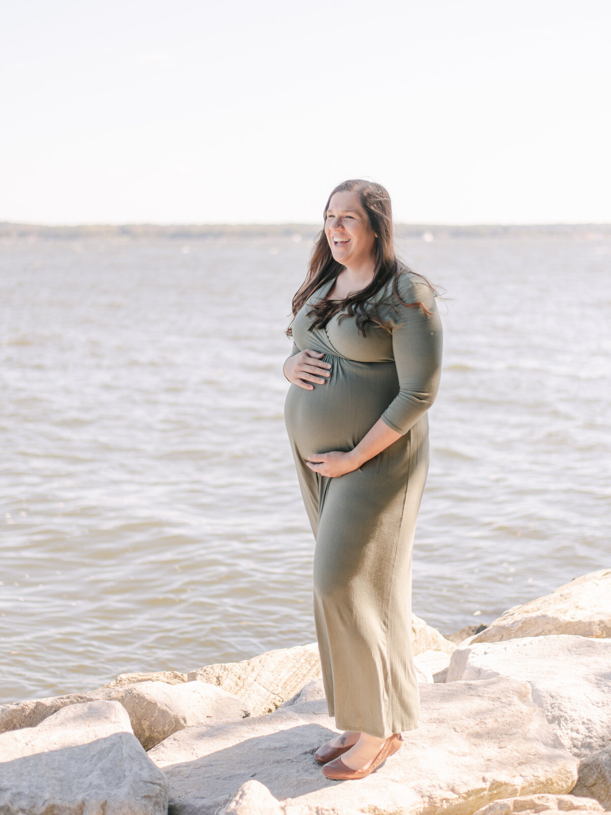 Mom holding her pregnant belly while standing on rocks near beach in Virginia.