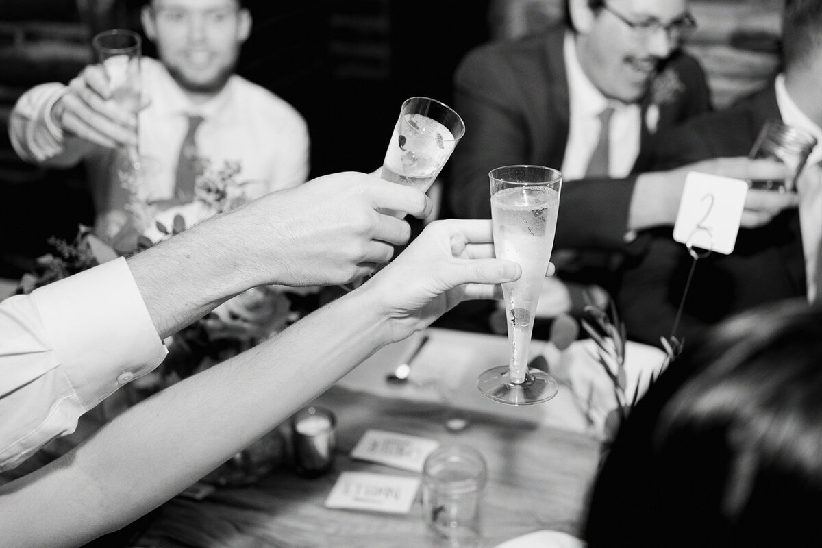 A black and white documentary-style photo of Amy and Julian raising celebratory drinks at their wedding in Bahama, North Carolina, captured by North Carolina wedding photographer Jessica Leigh Mason.