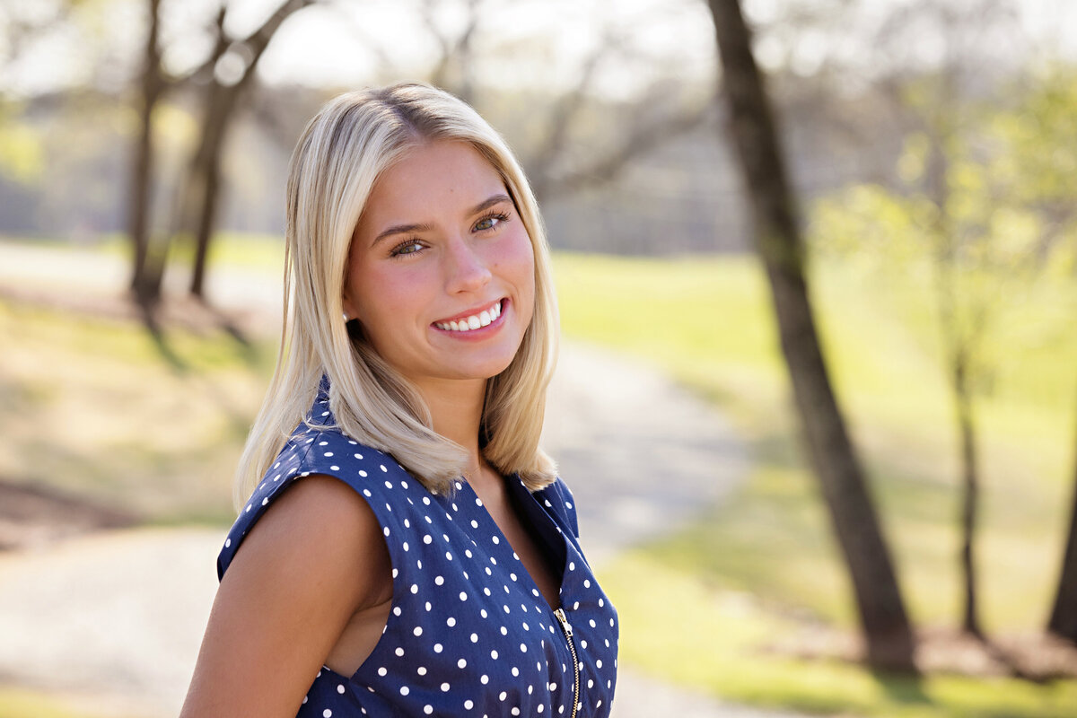 Close-up spring portrait of high school senior with blonde hair smiling in a blue polka dot dress, framed by soft greenery and a nature trail in Charlotte NC