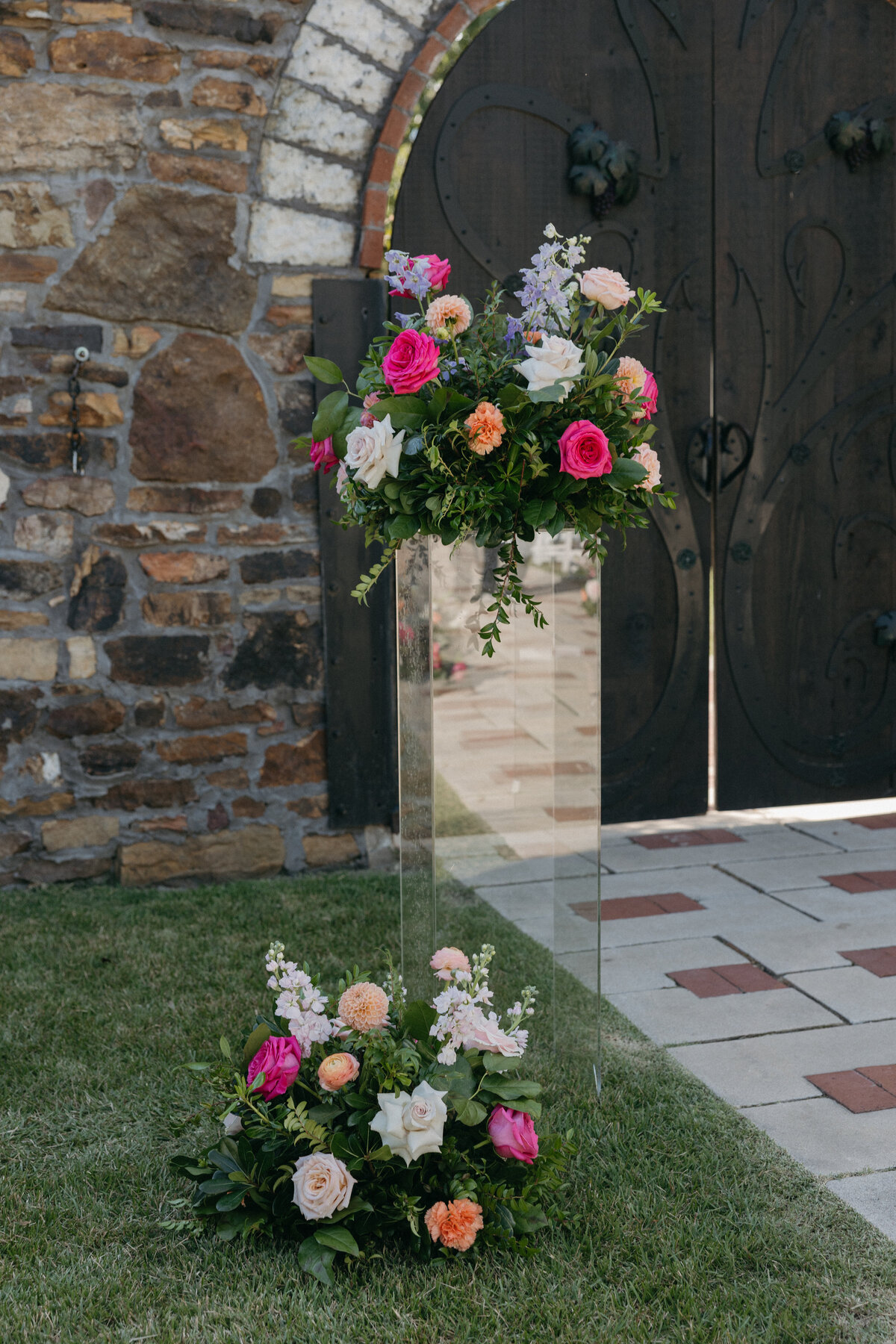 Tall wedding ceremony floral installation with bright garden roses, dahlias, and greenery on a clear pedestal, displayed against a stone chapel backdrop.
