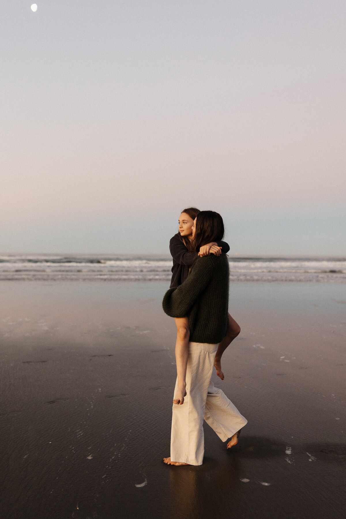 A mother carrying her daughter and walking along the beach at a family photoshoot during sunset with lucy jane photography in christchurch