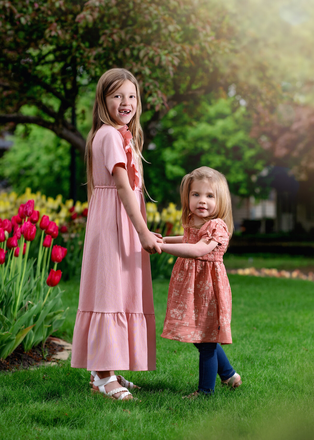 Two young sisters are holding hands and smiling in a vibrant garden with red tulips and green grass.