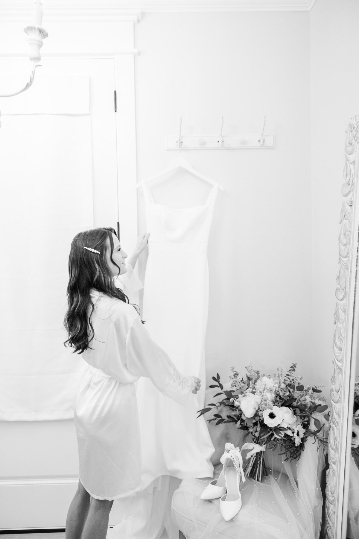A artistic black and white image of a bride standing with her wedding dress as she looks at it hanging on the wall.
