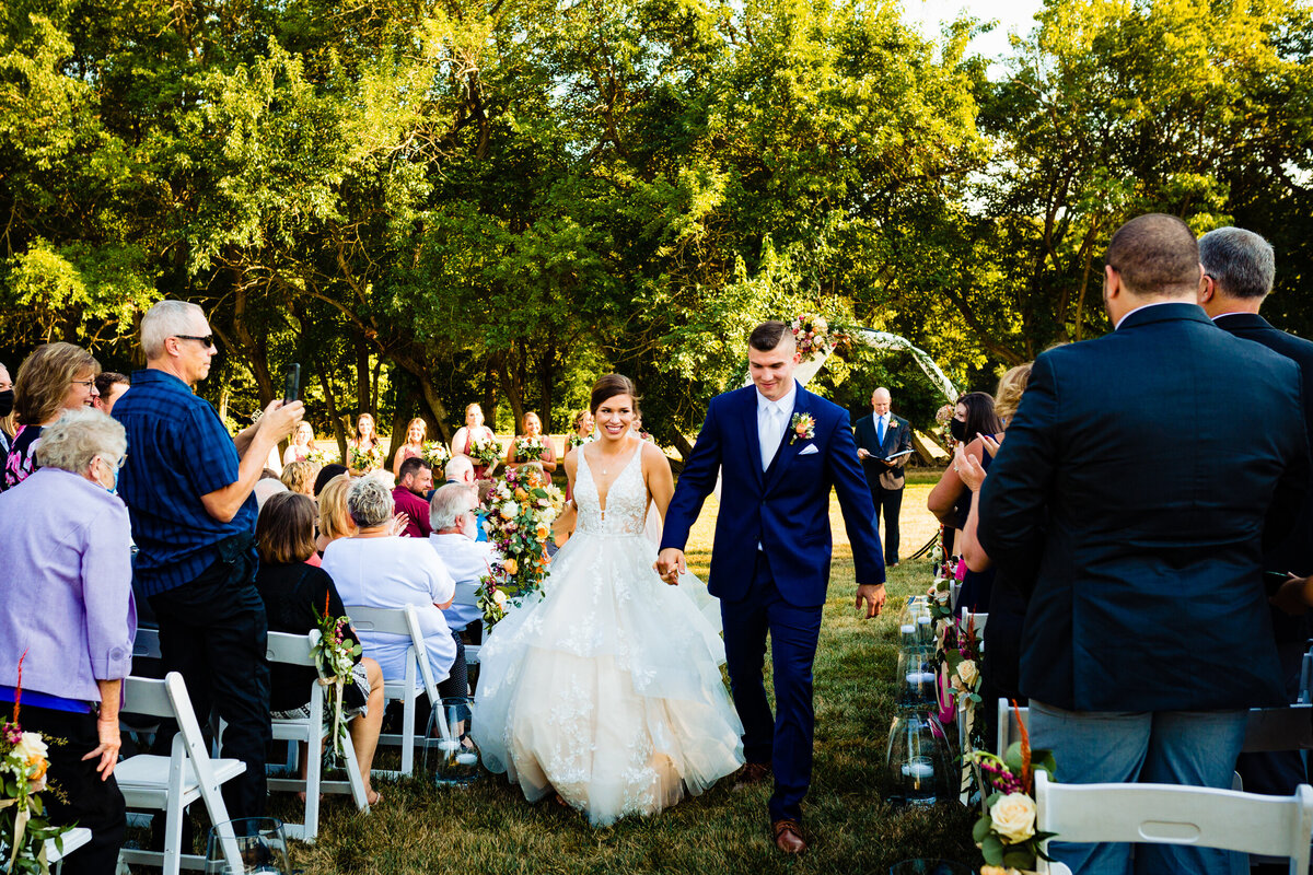 One of the top wedding photos of 2020. Taken by Adore Wedding Photography- Toledo, Ohio Wedding Photographers. This photo is of a bride and groom smiling and holding hands walking down the aisle after getting married outside at Nazareth Hall in Grand Rapids