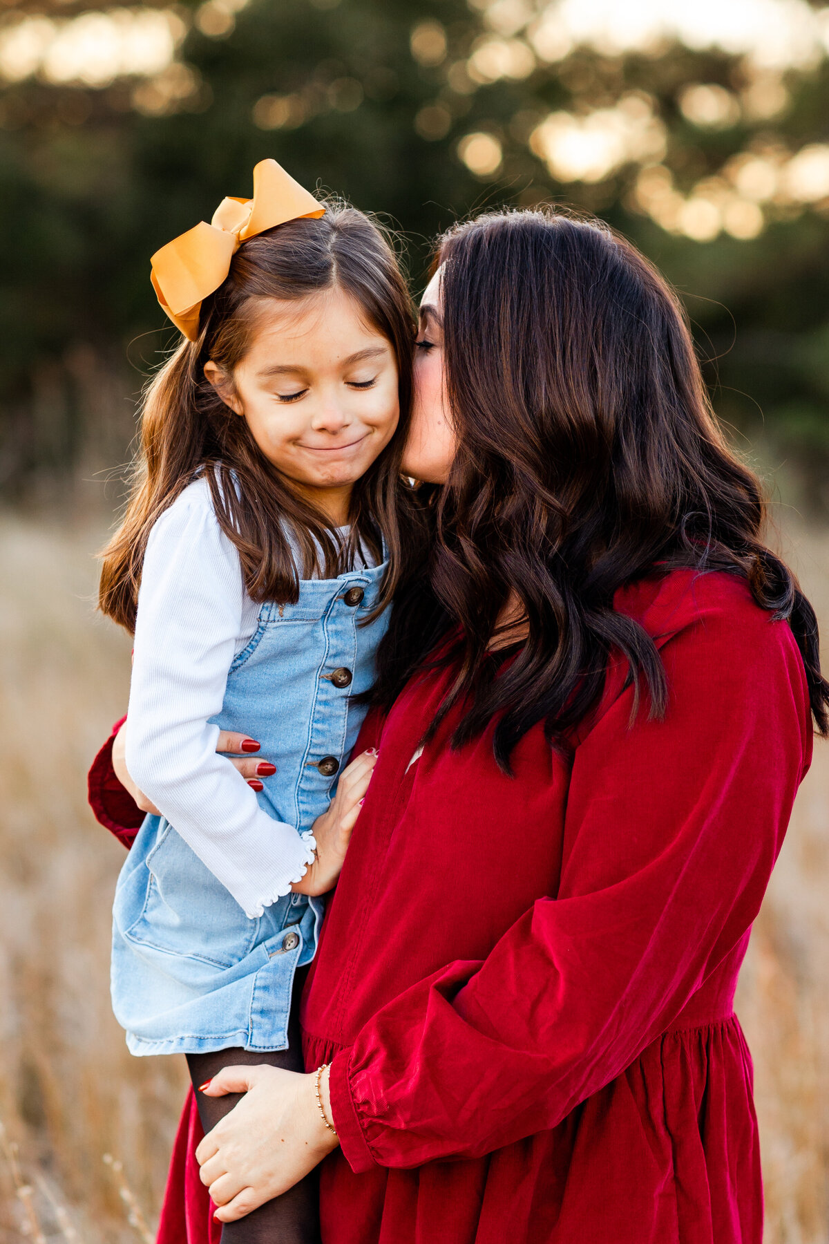 A mother in a red dress kisses her young daughters cheek during a cozy fall session in Virginia.  The little girl is smiling in a denim overall dress as golden light filters through the trees.  