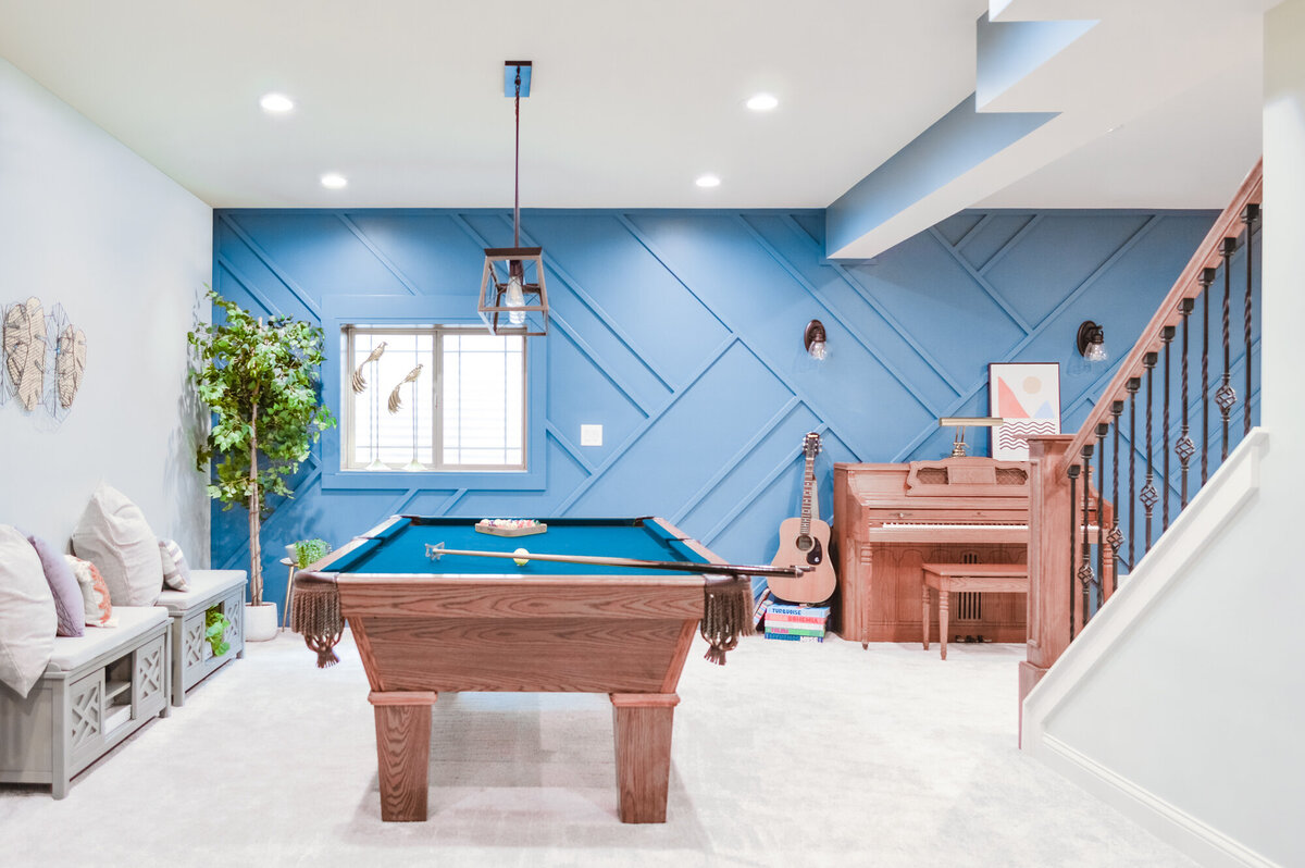 A spacious recreational room with light blue walls featuring a striking geometric paneling design. A wooden pool table with a teal felt surface is centrally located on a light gray carpet, with a cue stick resting on its side. Above the table, a modern pendant light with a wooden frame and exposed bulb hangs from the ceiling. To the right of the pool table, an upright piano in a light wood finish stands against the wall, flanked by an acoustic guitar. A framed abstract print rests on top of the piano. To the left, a long gray bench with white cushions and storage compartments sits against the wall, adorned with decorative pillows and a potted artificial tree. A staircase with dark wood railings and white risers is visible on the right, leading to another level. The room is well-lit by recessed ceiling lights and natural light from a window with blinds.