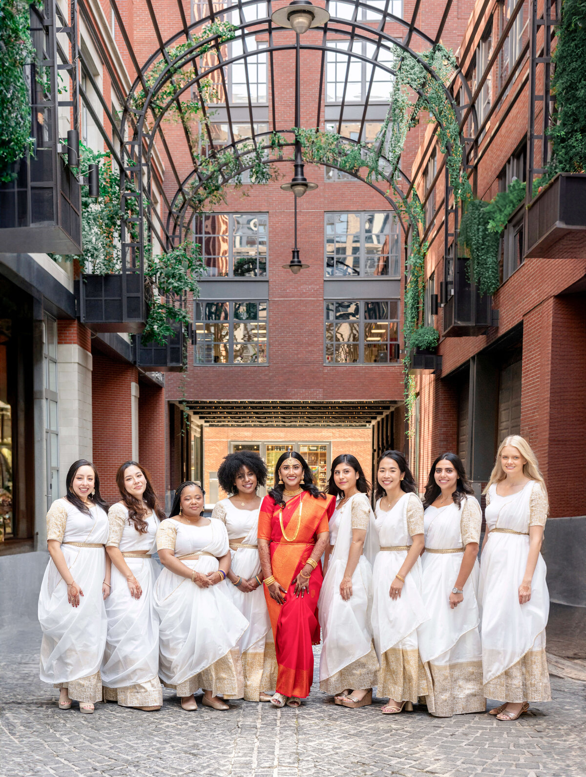 A group of women in traditional attire stands in an urban setting. One woman in a red and gold saree is surrounded by others in white dresses with gold details. They are positioned under an arched structure with green plants, on a cobblestone path.