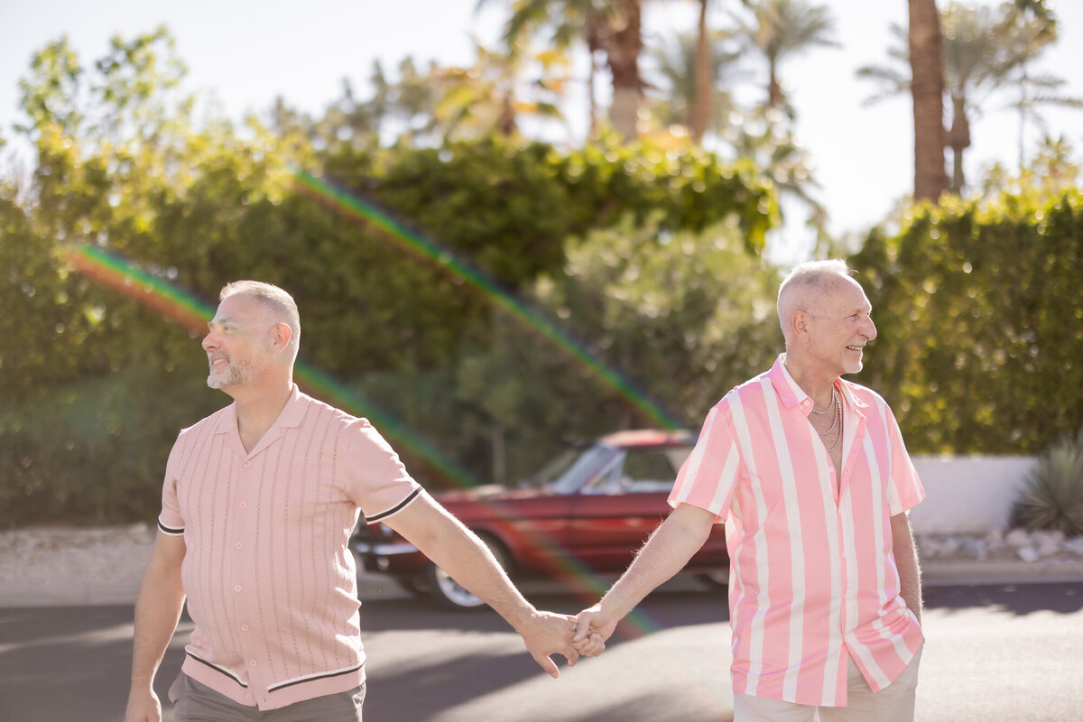 Couple walking hand in hand across the road, smiling and connected, framed by desert trees and classic car vibes.