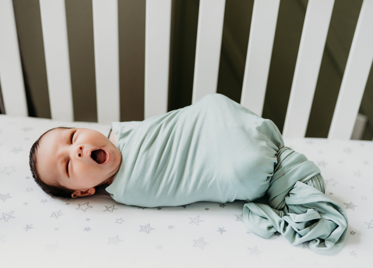 Newborn Photographer, a baby lays swaddled in a crib yawning