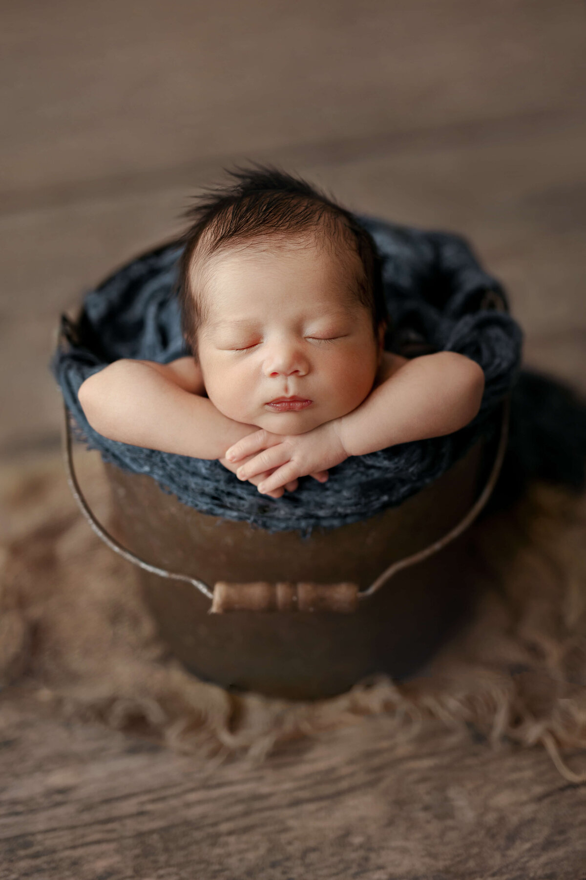 newborn baby boy laying in a metal bucket with a blue wrap on it with his head on this hands on a wooden floor in a newborn session