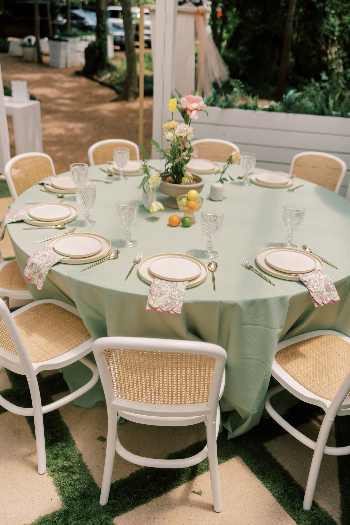 Outdoor round table set with sage green linen, white cane-back chairs, gold flatware, patterned napkins, and a citrus floral centerpiece.