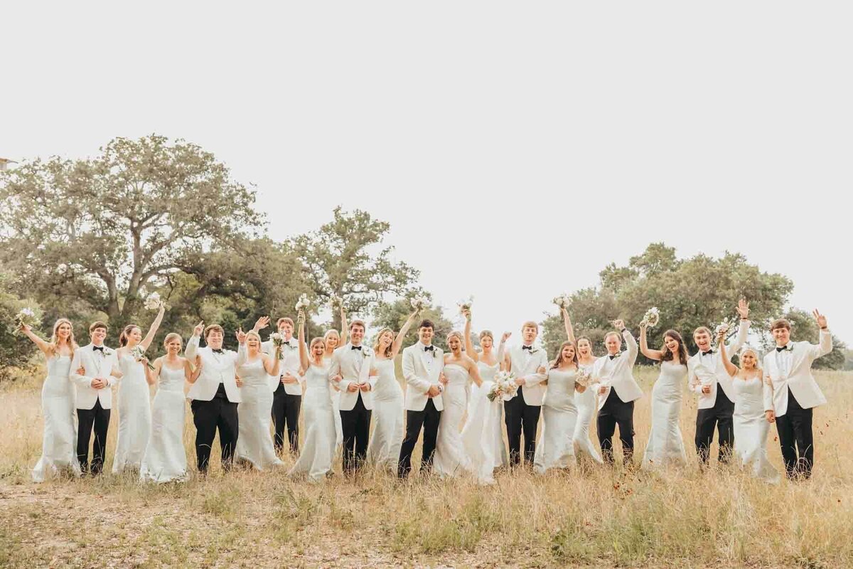 bride and groom celebrate with their hands in the air with their entire wedding party, in a field in roundtop texas.