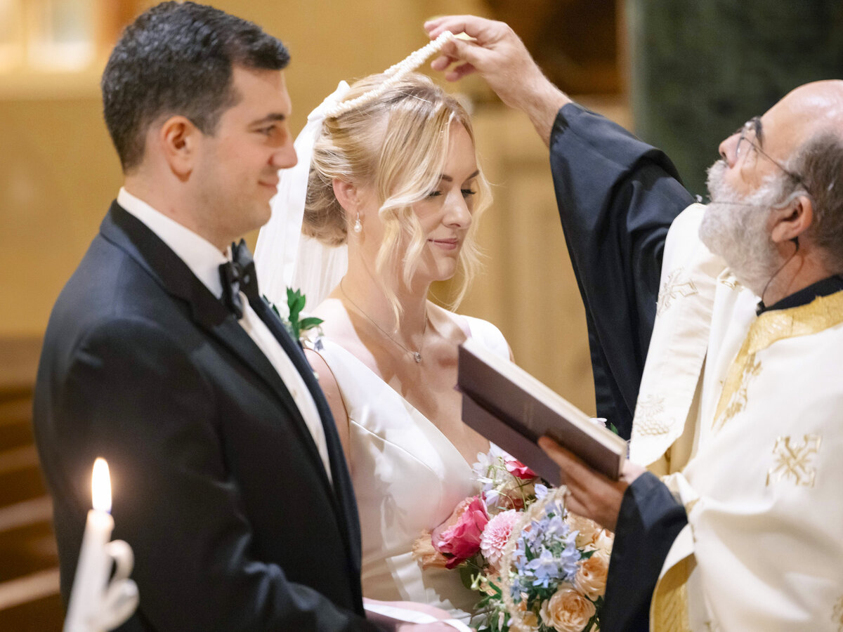 A bride and groom stand side by side in a church, holding a bouquet. A priest in ceremonial robes places a delicate crown on the brides head. Candles are visible in the foreground, creating a warm and intimate atmosphere.