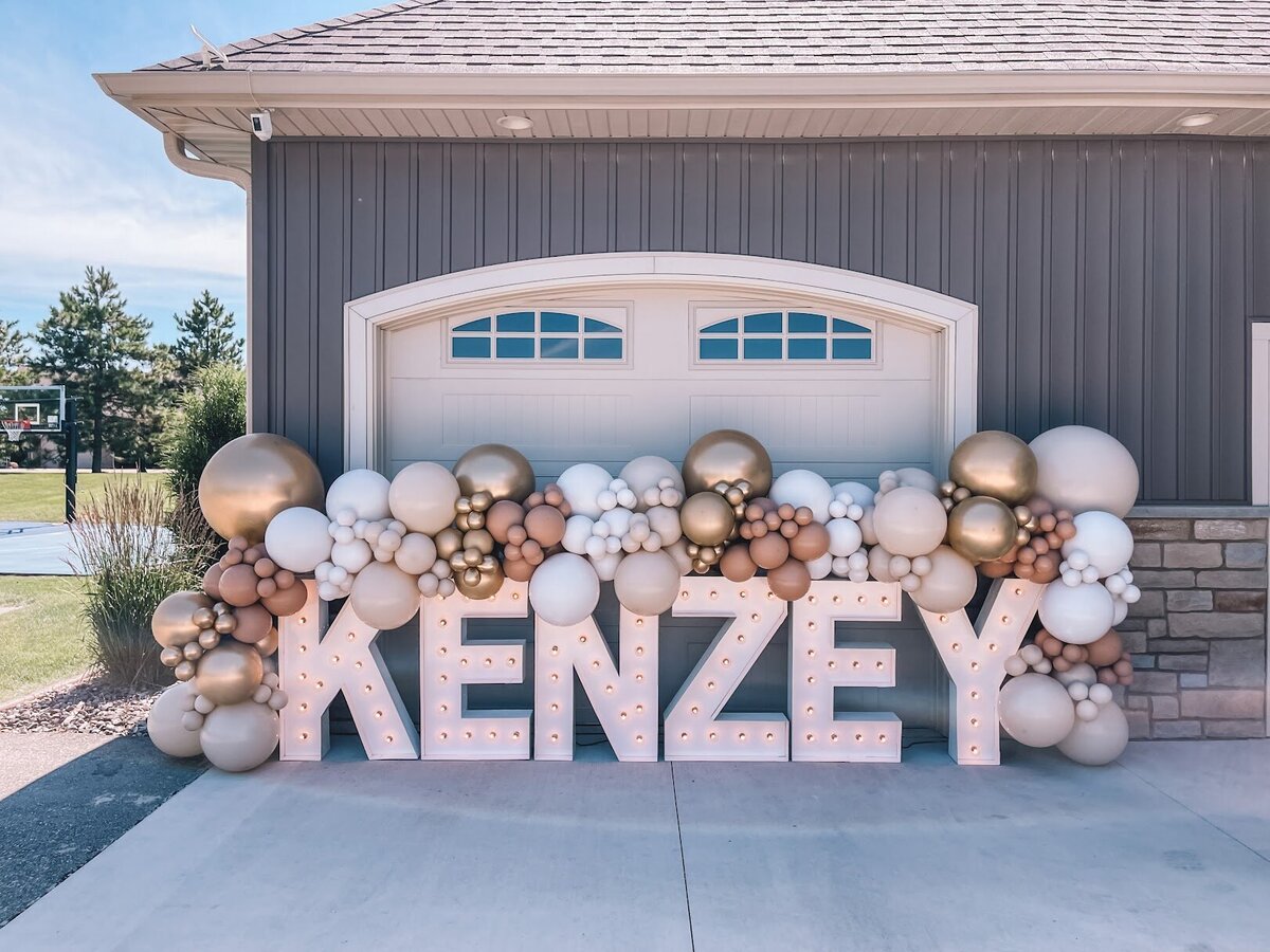 Gold, white, and tan balloons attached to giant marquee letters that spell the name Kenzey.