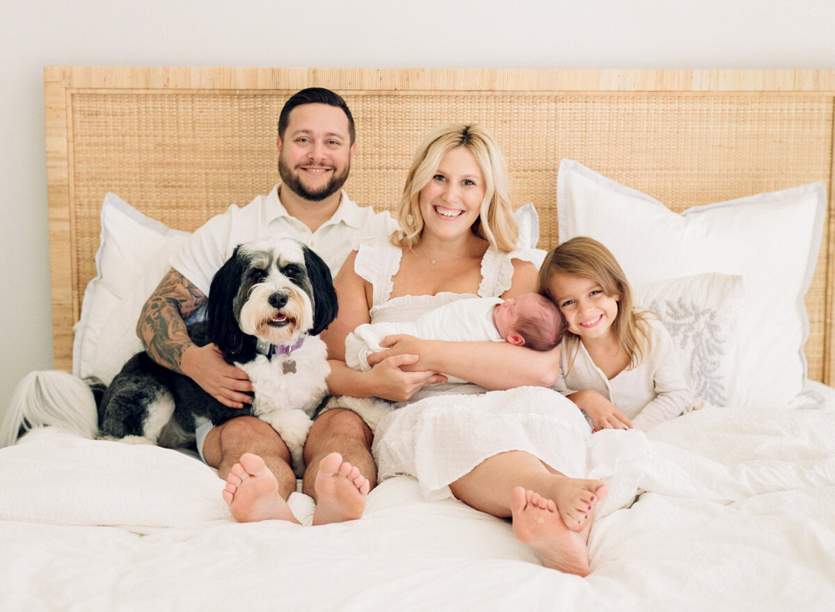 Family poses for a portrait with their bernadoodle while sitting on their bed. Image by Raleigh newborn photographer A.J. Dunlap Photography.