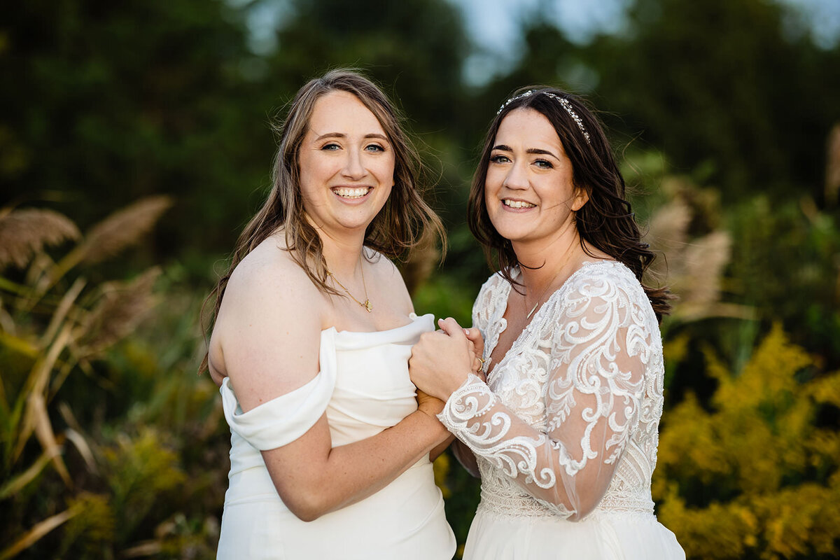 Two brides standing together amidst tall grass, one in a white off-the-shoulder dress and the other in a lace gown, both smiling at the camera.
