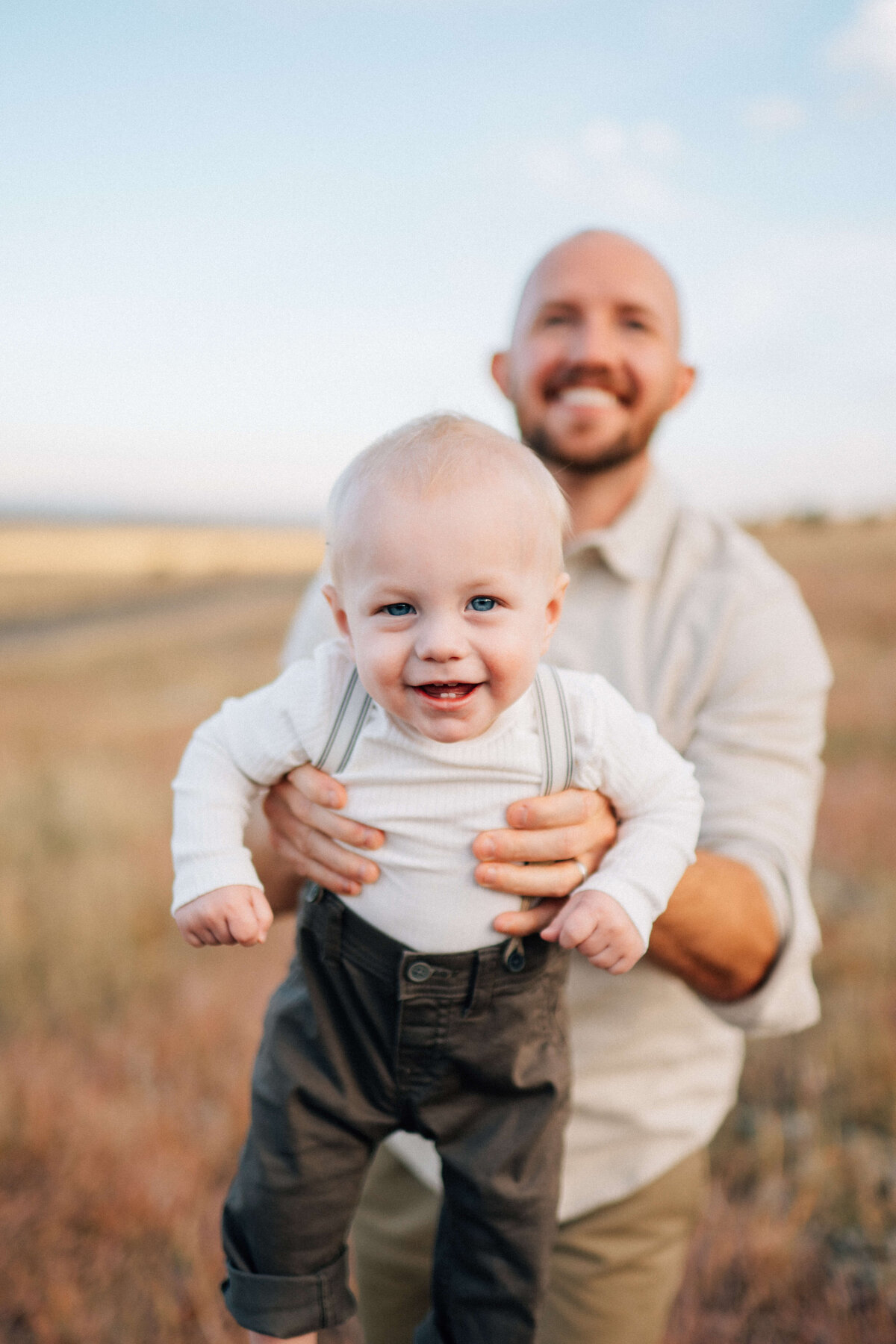 Dad holding his one year old boy in suspenders out toward the photographer while they smile