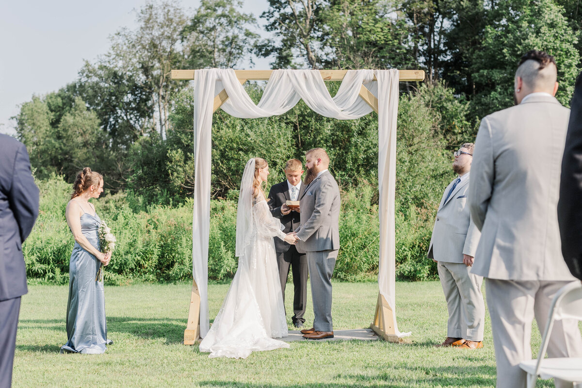 bride and groom holding hands under ceremony arch with bridal party and guests during wedding photo session in upstate ny 
