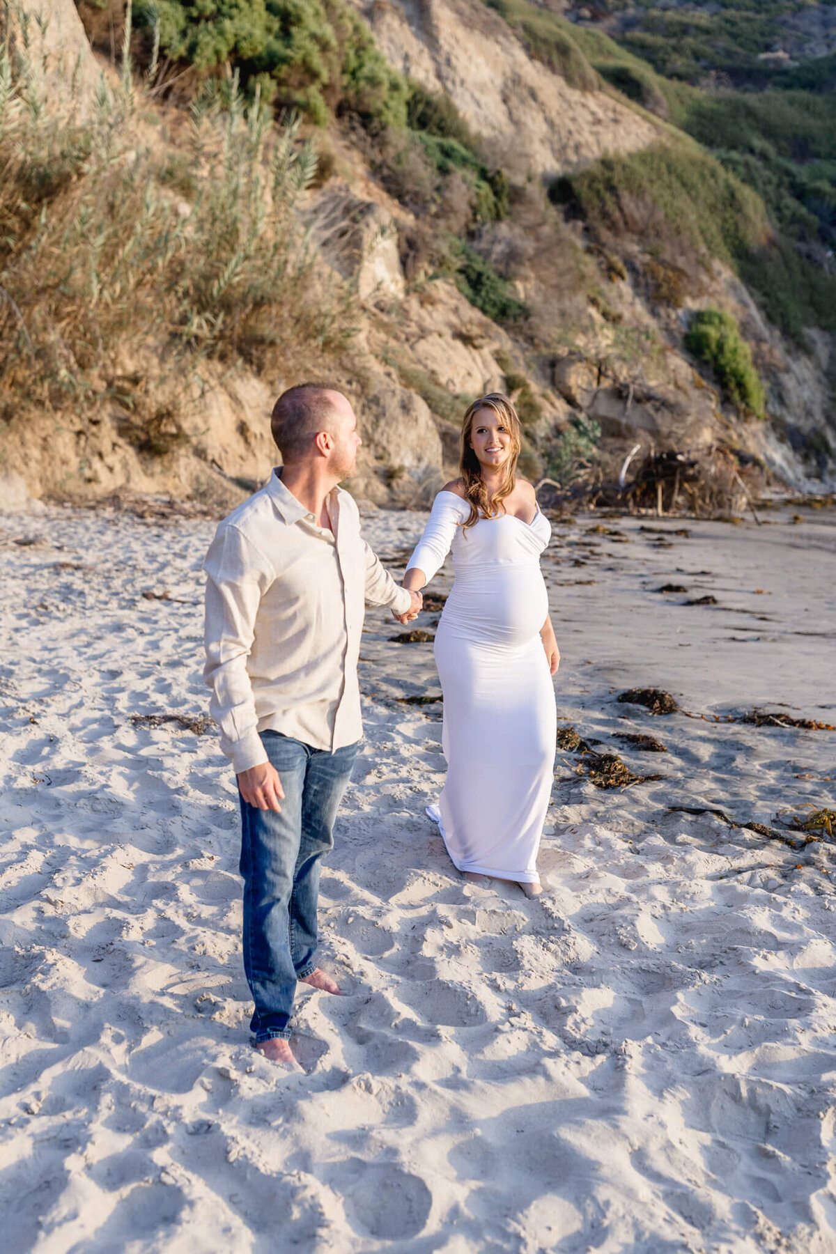 A couple stands on a sandy beach near rocky cliffs. The woman, wearing a long white dress, appears pregnant and holds hands with the man, who is wearing a beige shirt and jeans. The scene is set in soft sunlight.