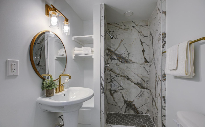 Modern bathroom remodel featuring a walk-in marble-tiled shower with hexagon floor tile, pedestal sink with gold fixtures, and built-in shelving—combining style and smart use of space.